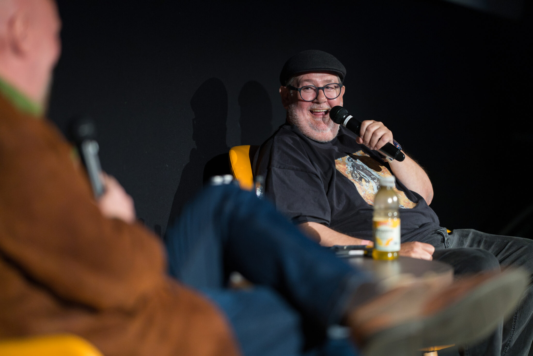 Two people sit and talk on stage. One man, wearing glasses and a black cap, smiles and speaks into a microphone. The other person is blurred in the foreground. A drink bottle is on the table in front of them.