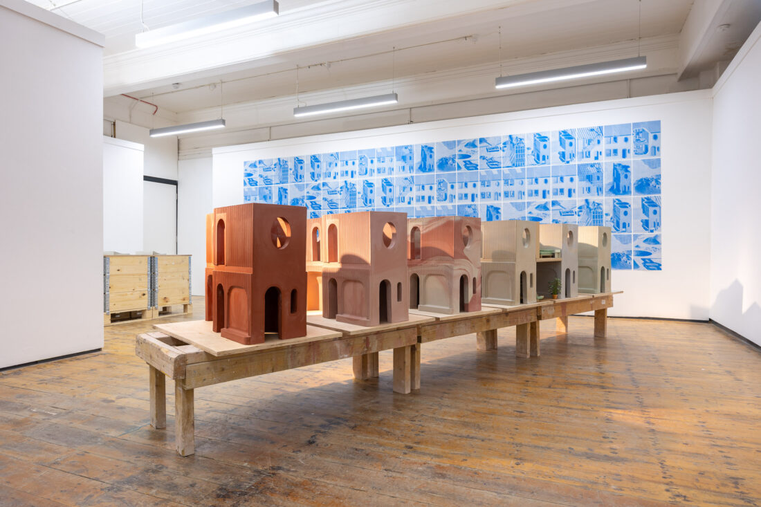 A row of architectural models sits on a wooden table in a gallery with white walls and wooden floors. A large blue and white mural with building sketches is displayed on the wall behind the models.