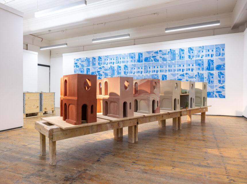 A row of architectural models sits on a wooden table in a gallery with white walls and wooden floors. A large blue and white mural with building sketches is displayed on the wall behind the models.
