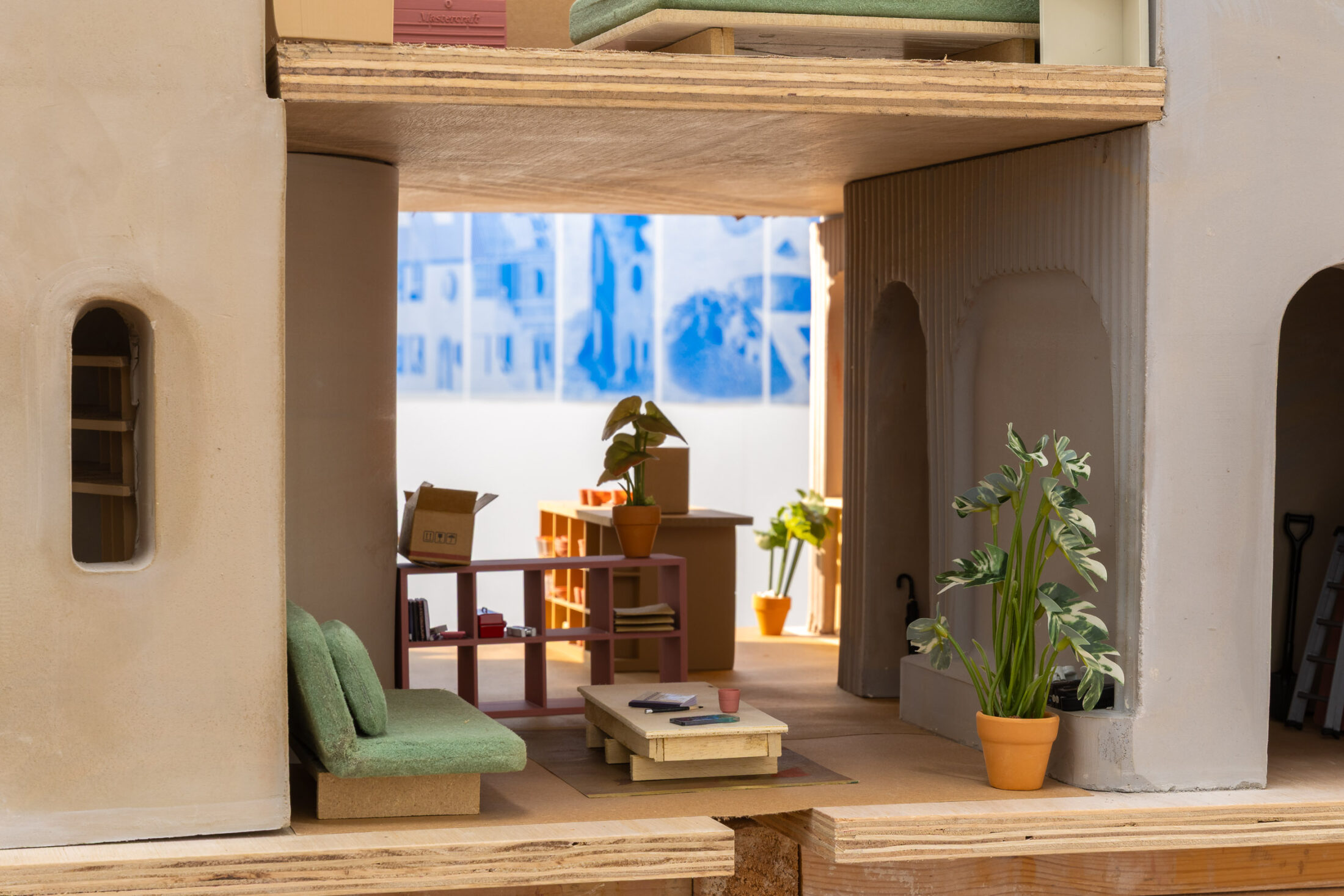 A miniature modern living room and kitchen with green sofas, potted plants, a coffee table, and cardboard boxes. Sunlight streams in through windows in the background.