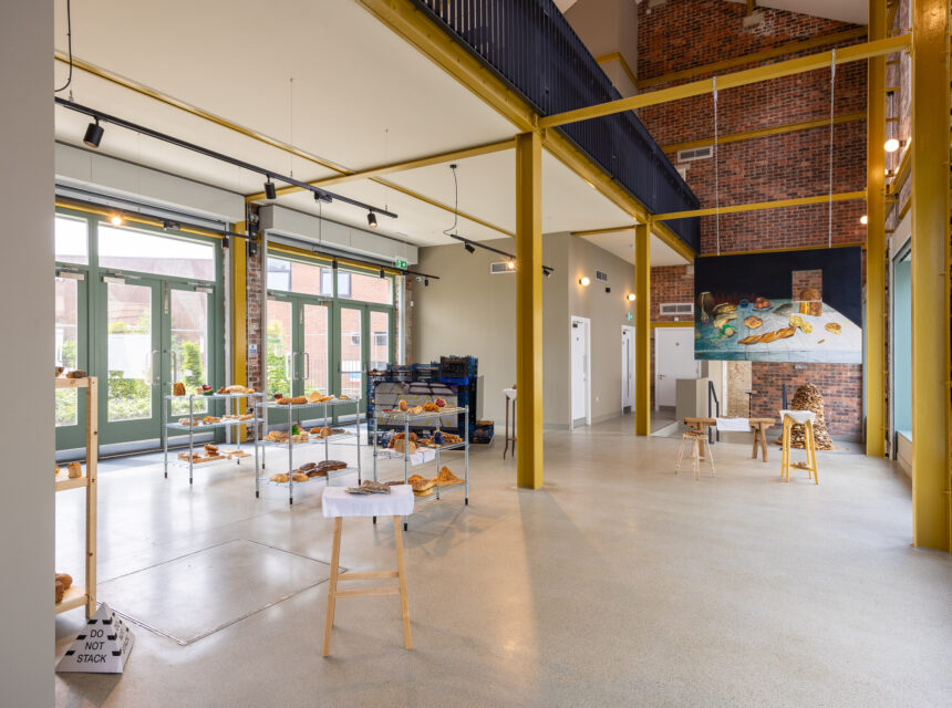 A spacious, modern room with large windows, yellow beams, and scattered tables displaying various breads. There are artworks on the walls and stacked trays near the back.