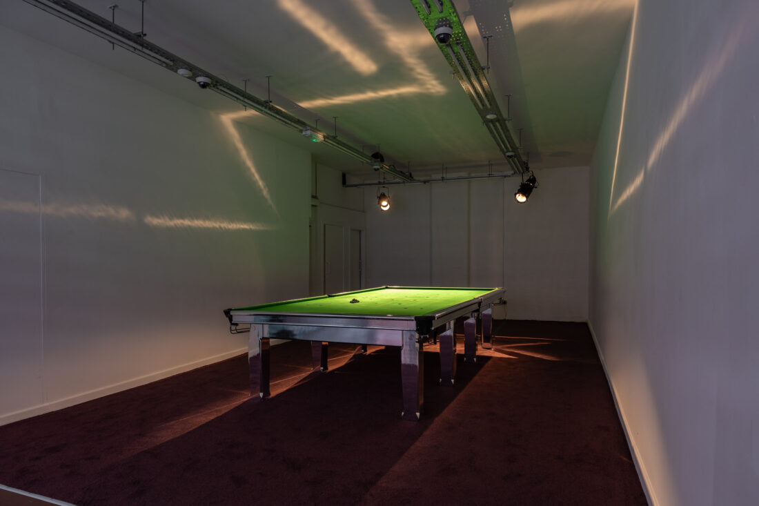 A snooker table with a green surface is centred in a dimly lit, plain room with cream walls and a dark carpet. Overhead spotlights cast shadows on the walls and ceiling.