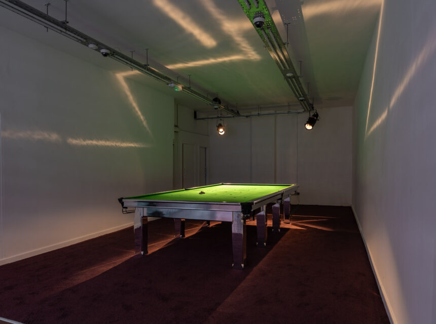 A snooker table with a green surface is centred in a dimly lit, plain room with cream walls and a dark carpet. Overhead spotlights cast shadows on the walls and ceiling.