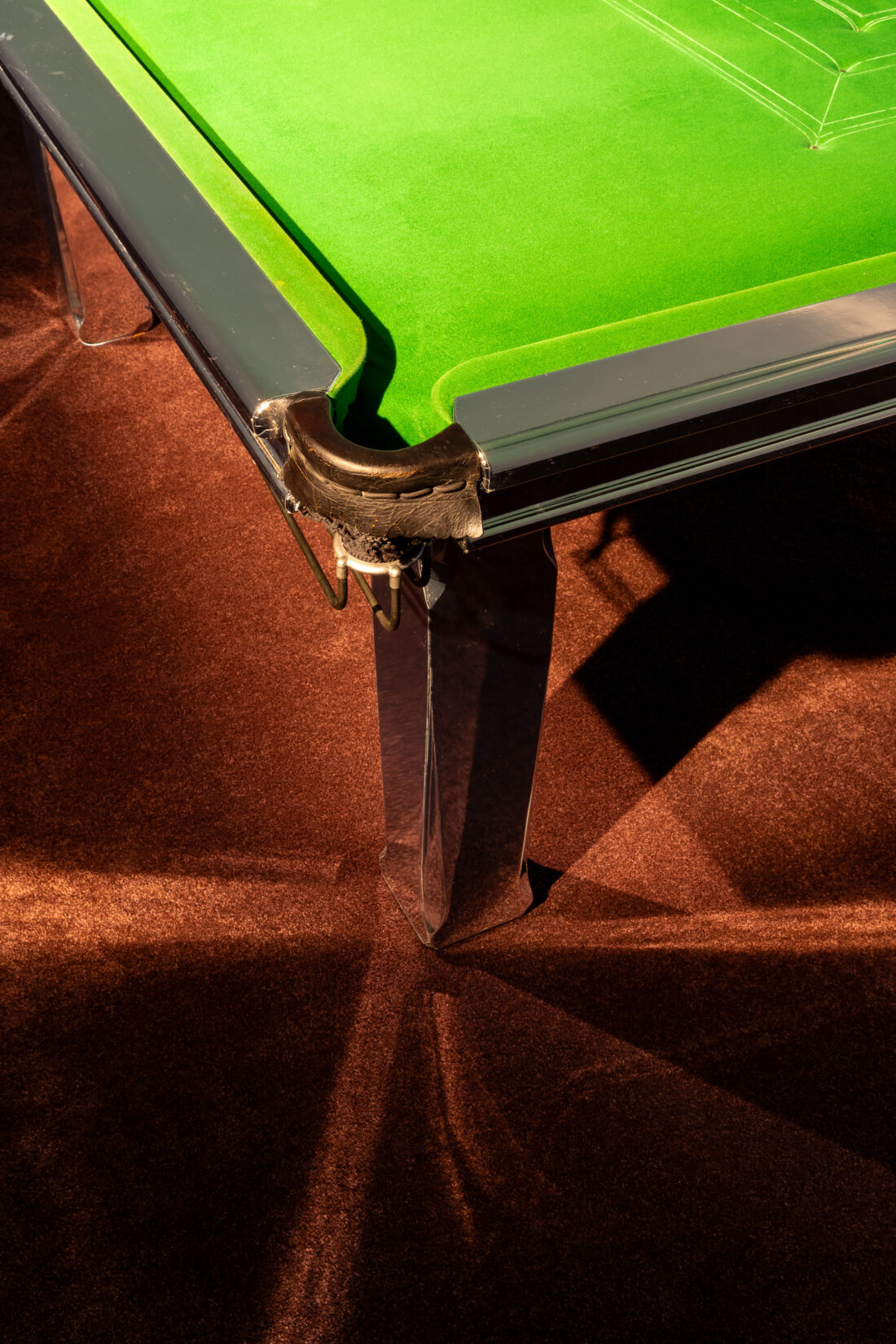 The image shows a close-up of a snooker or pool table corner with green baize, a metal pocket, and brown carpet underneath, illuminated by warm lighting.