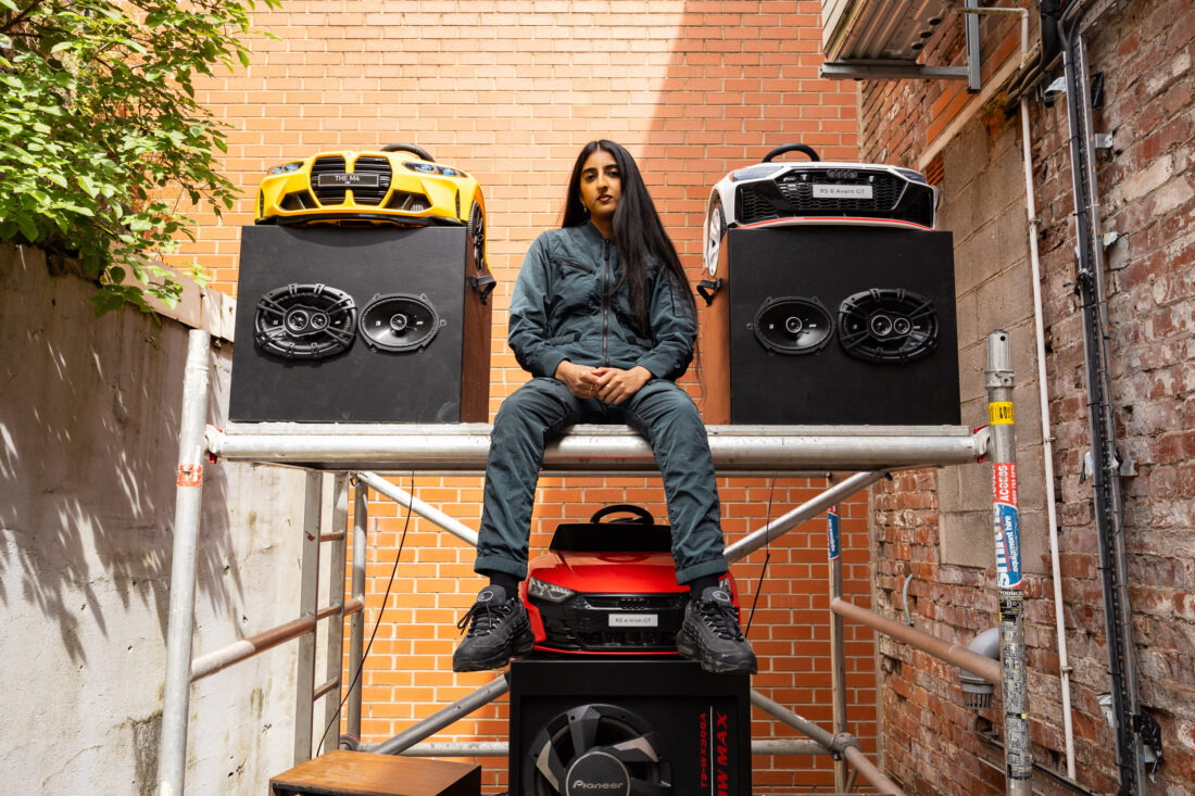 A person in dark clothing sits on scaffolding between two large speakers with toy car fronts attached, set against a brick wall in an alleyway.