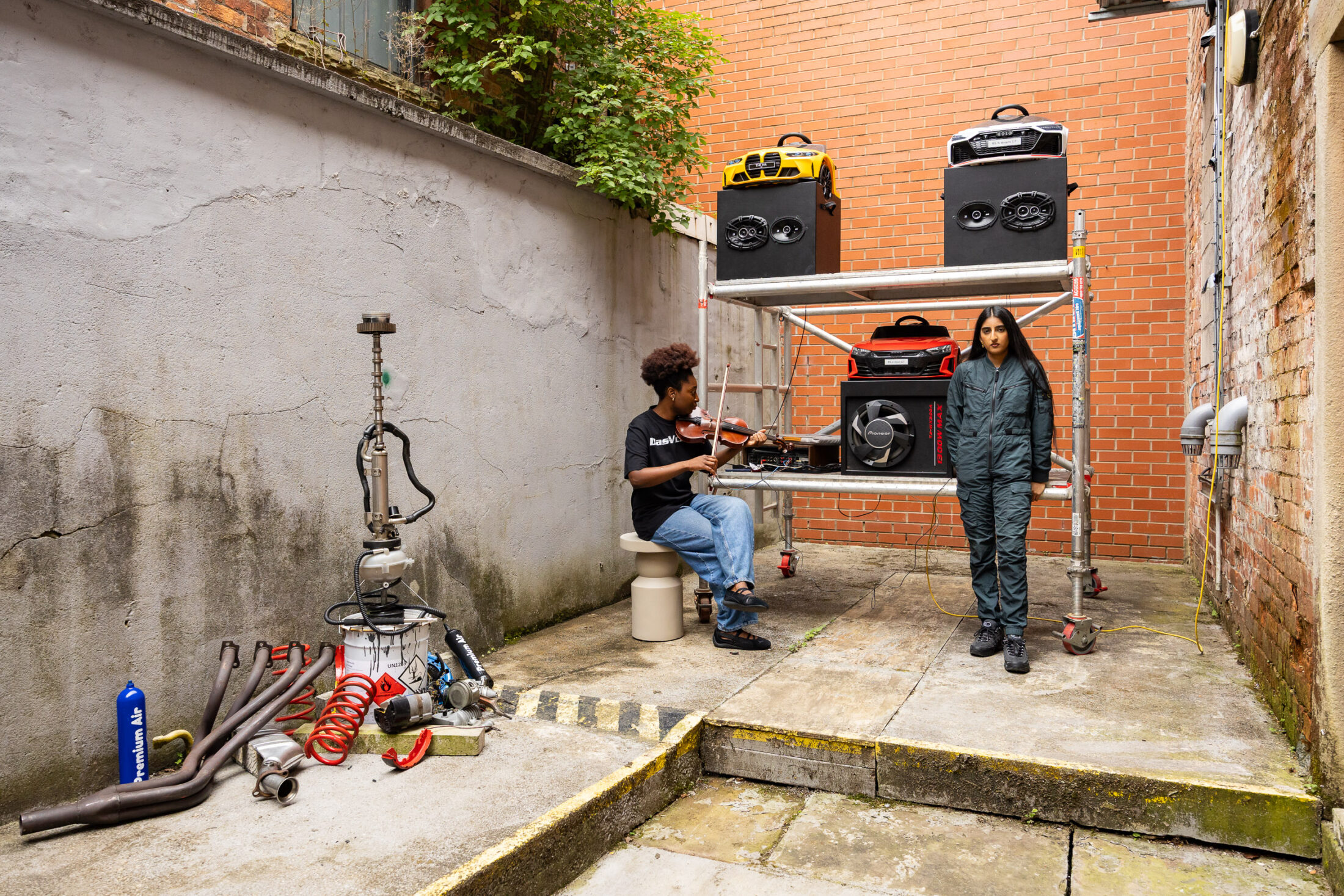 Two people are in an alley; one plays a violin while sitting, the other stands beside them. Behind them is scaffolding holding speakers and music equipment; assorted pipes and tools are scattered nearby.