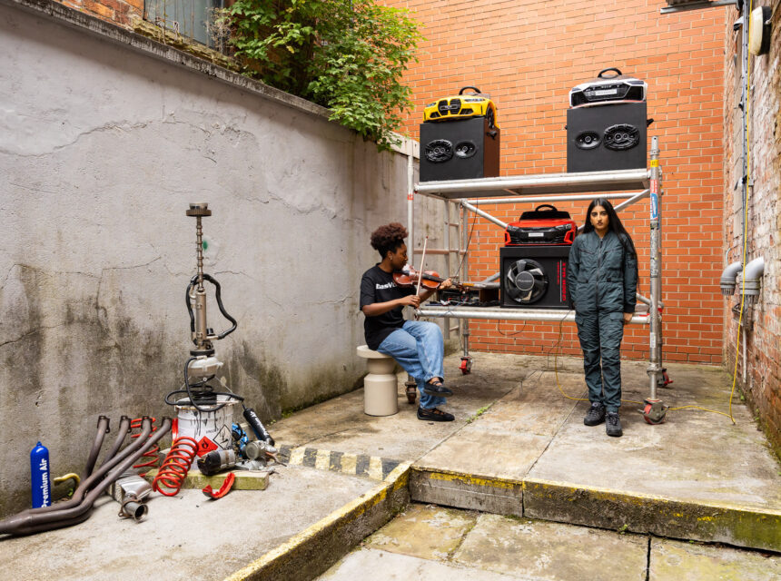 Two people in an alley: one sits playing a violin, the other stands beside scaffolding stacked with speakers and tools. Car parts and machinery are scattered on the ground nearby. Brick and concrete walls surround them.
