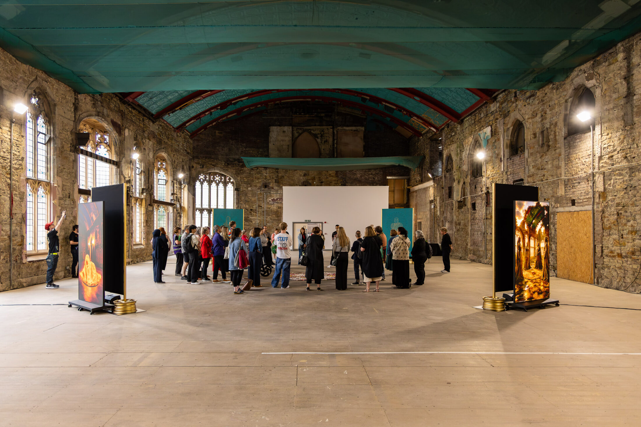 A group of people gathers in a large, rustic hall with stone walls, arched windows, and art displays on either side. The ceiling is teal, and the space is lit with natural and artificial light.