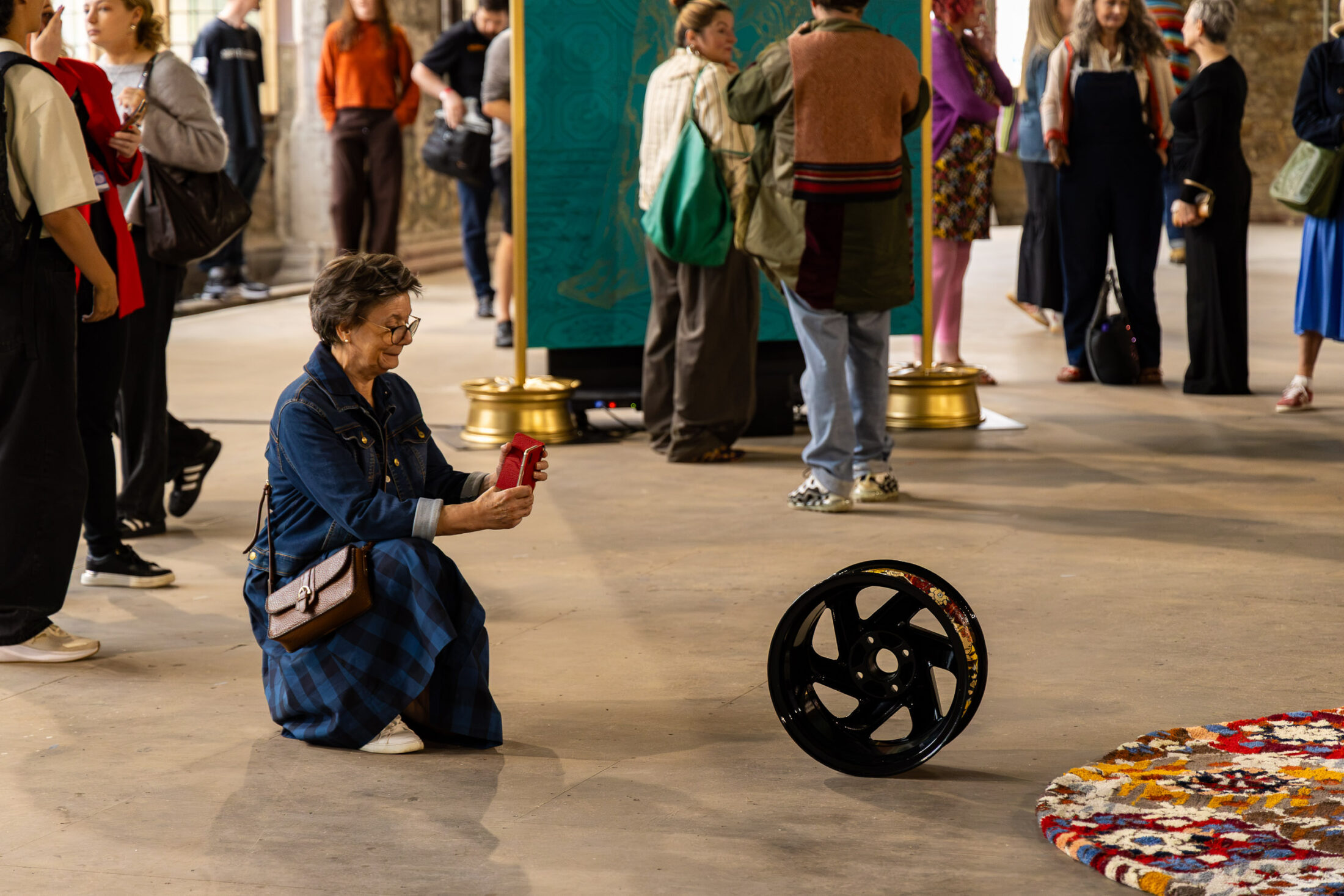 An woman kneels to take a photo of a black metal wheel displayed on the floor at an art exhibition, with other people and colourful artworks visible in the background.
