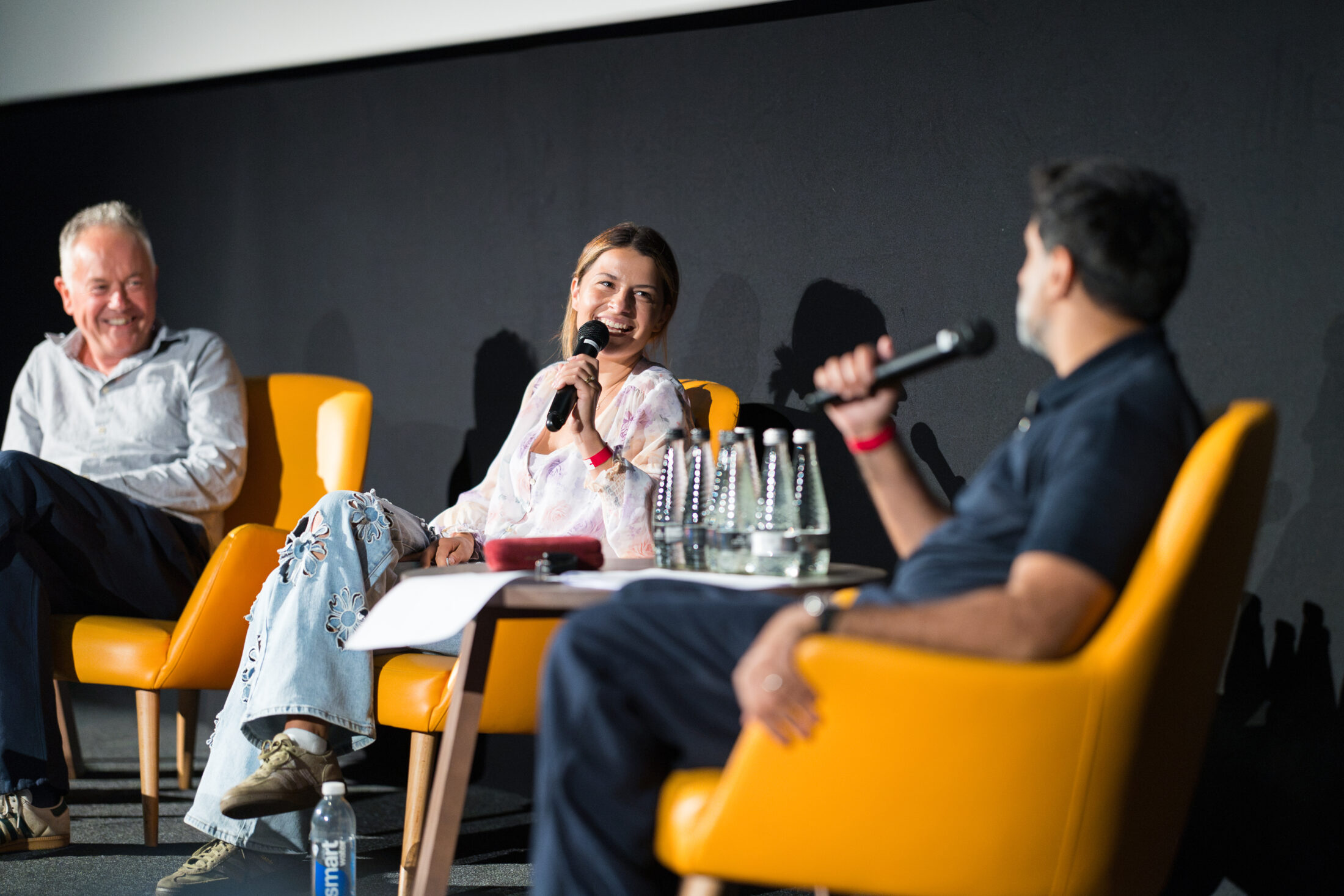 Three people sit in yellow chairs on stage, holding microphones and smiling during a panel discussion. A small table with water bottles is in front of them. The background is dark, suggesting an indoor event or conference.