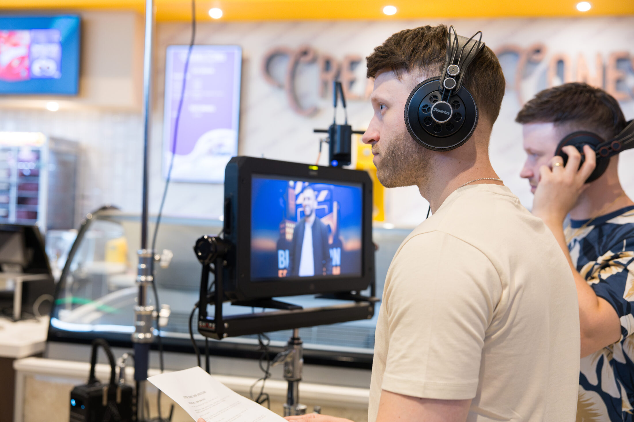 Two people wearing headphones stand near a monitor showing a person on screen, possibly filming or directing in a brightly lit indoor setting. One holds a paper script.