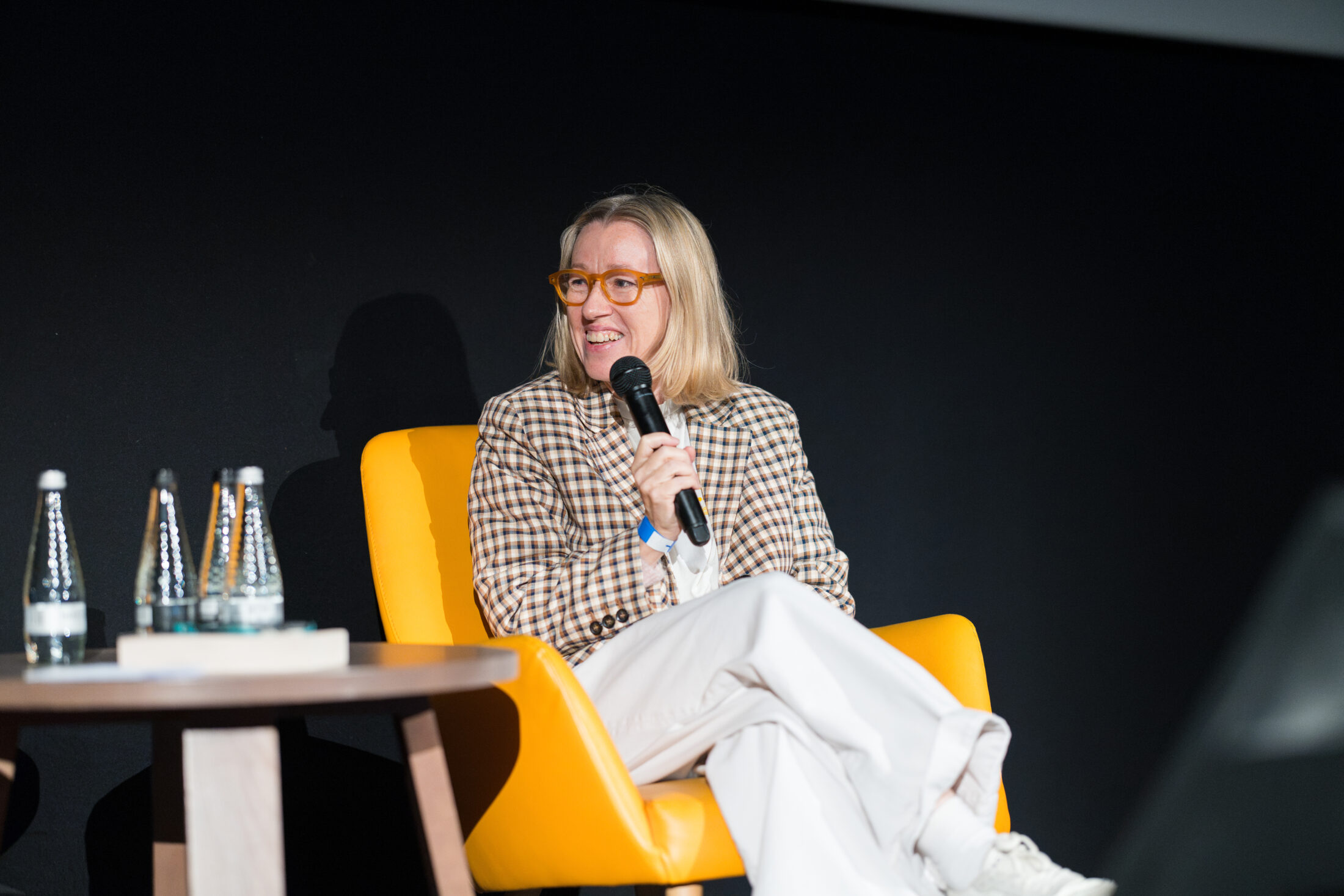 A person with blonde hair and glasses, wearing a checked blazer and white trousers, sits on a yellow chair holding a microphone. Two water bottles and a book are on a table nearby.