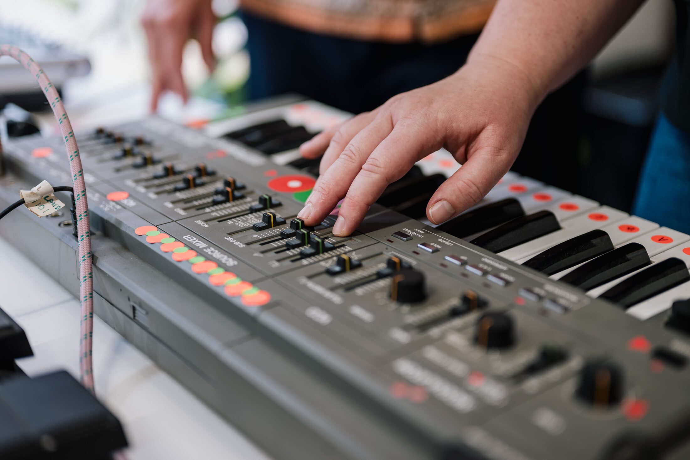 A close-up of a person adjusting sliders and playing keys on an electronic keyboard or synthesiser, with colourful buttons and controls visible.