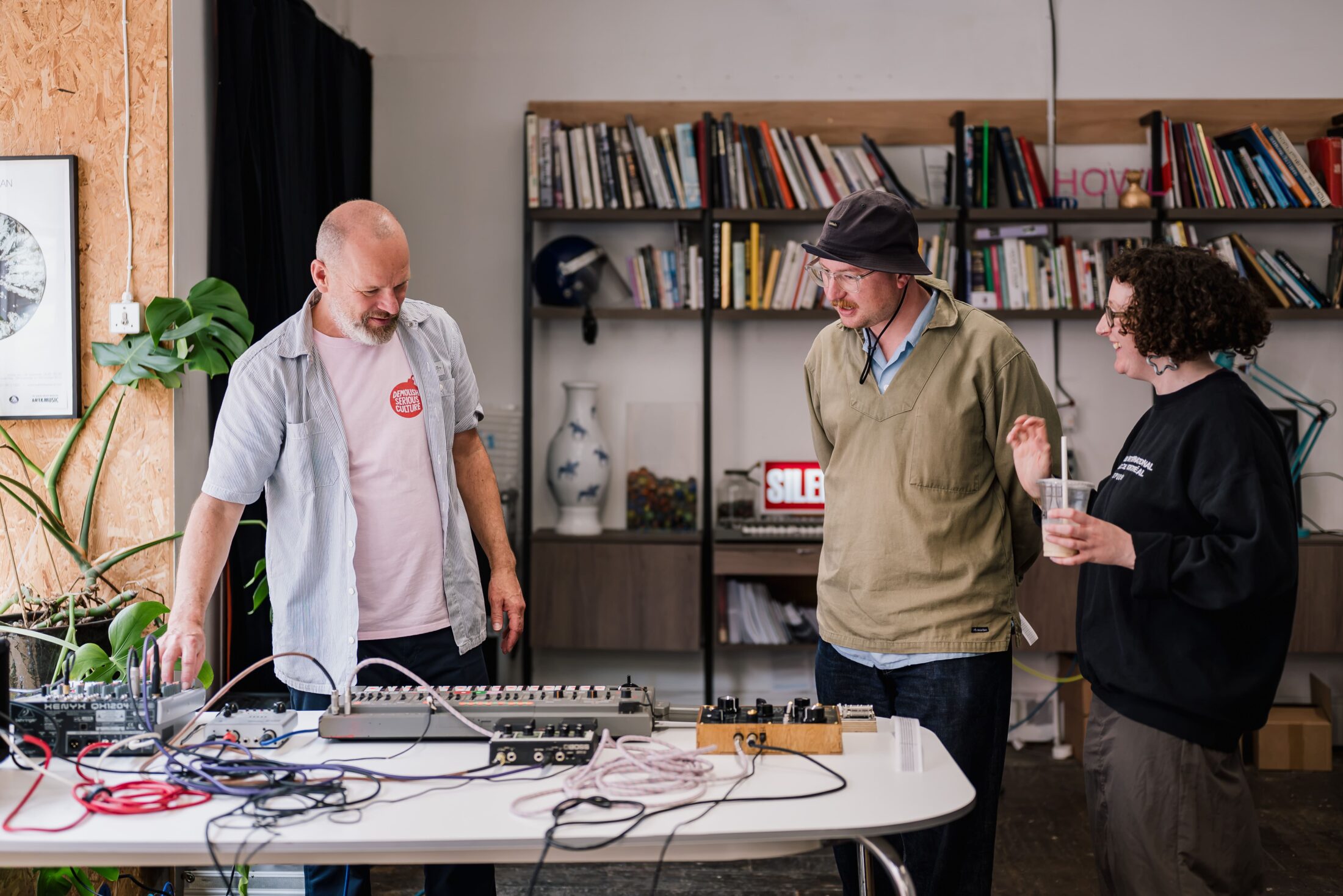 Three people stand around a table with electronic music equipment in a room with bookshelves. One person adjusts the equipment while the others watch and chat, one holding a drink.