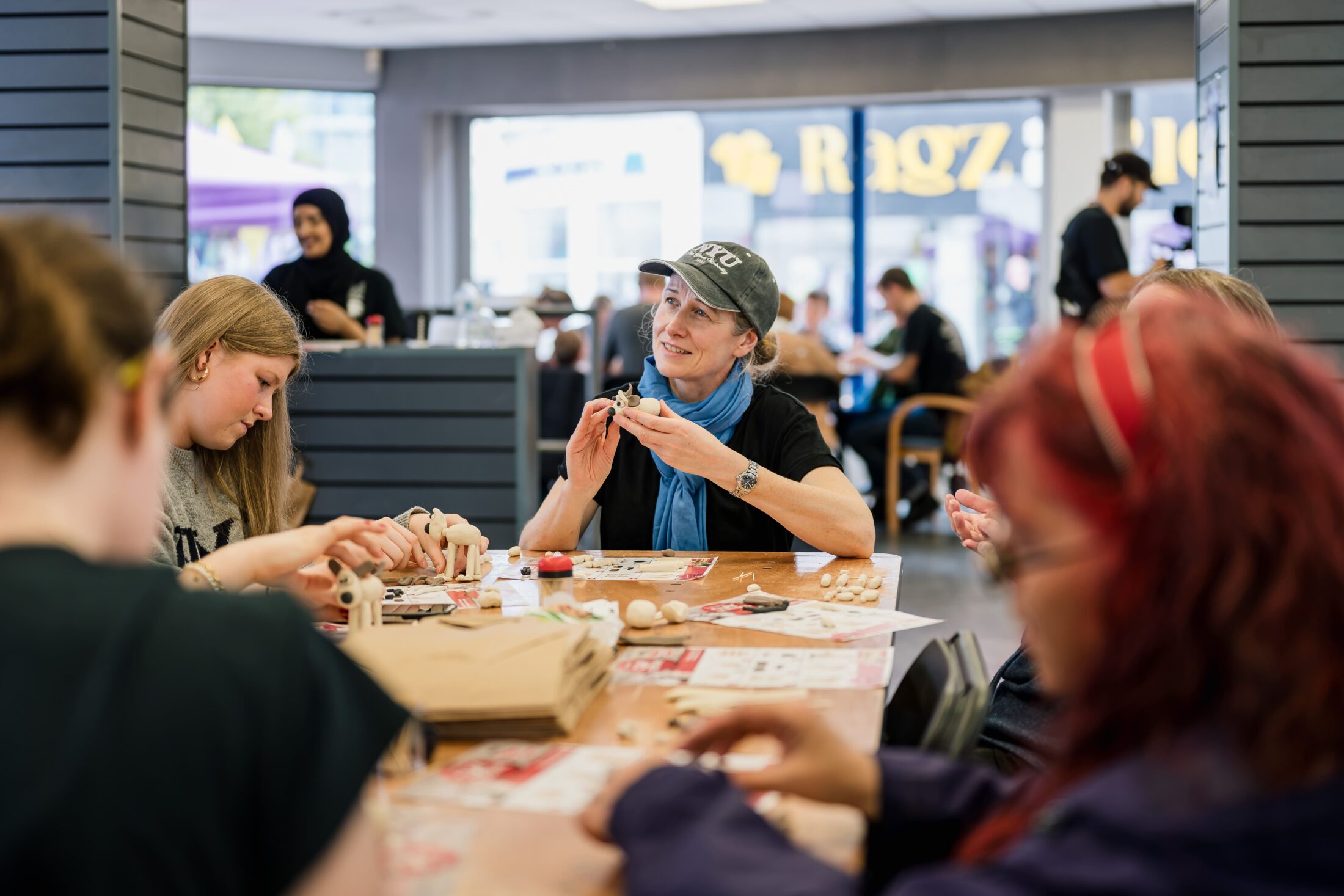 People sit around a table working on crafts in a bright room. A woman in a cap smiles whilst holding a small object. Others focus on projects. The background shows windows, people, and a sign reading Ragzi..