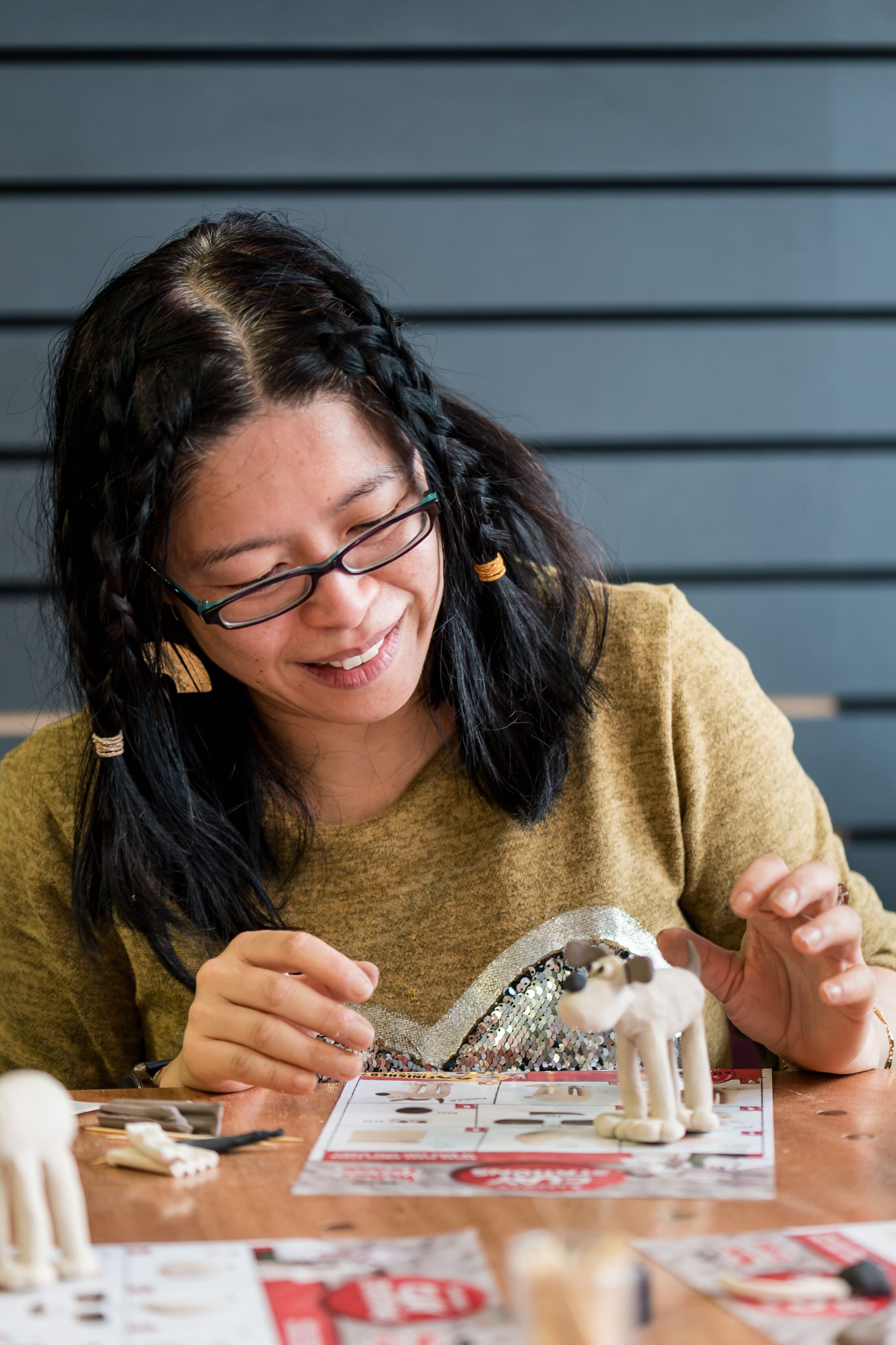 A woman with glasses and plaited hair smiles while assembling a small dog figurine at a table with instructions and craft materials.