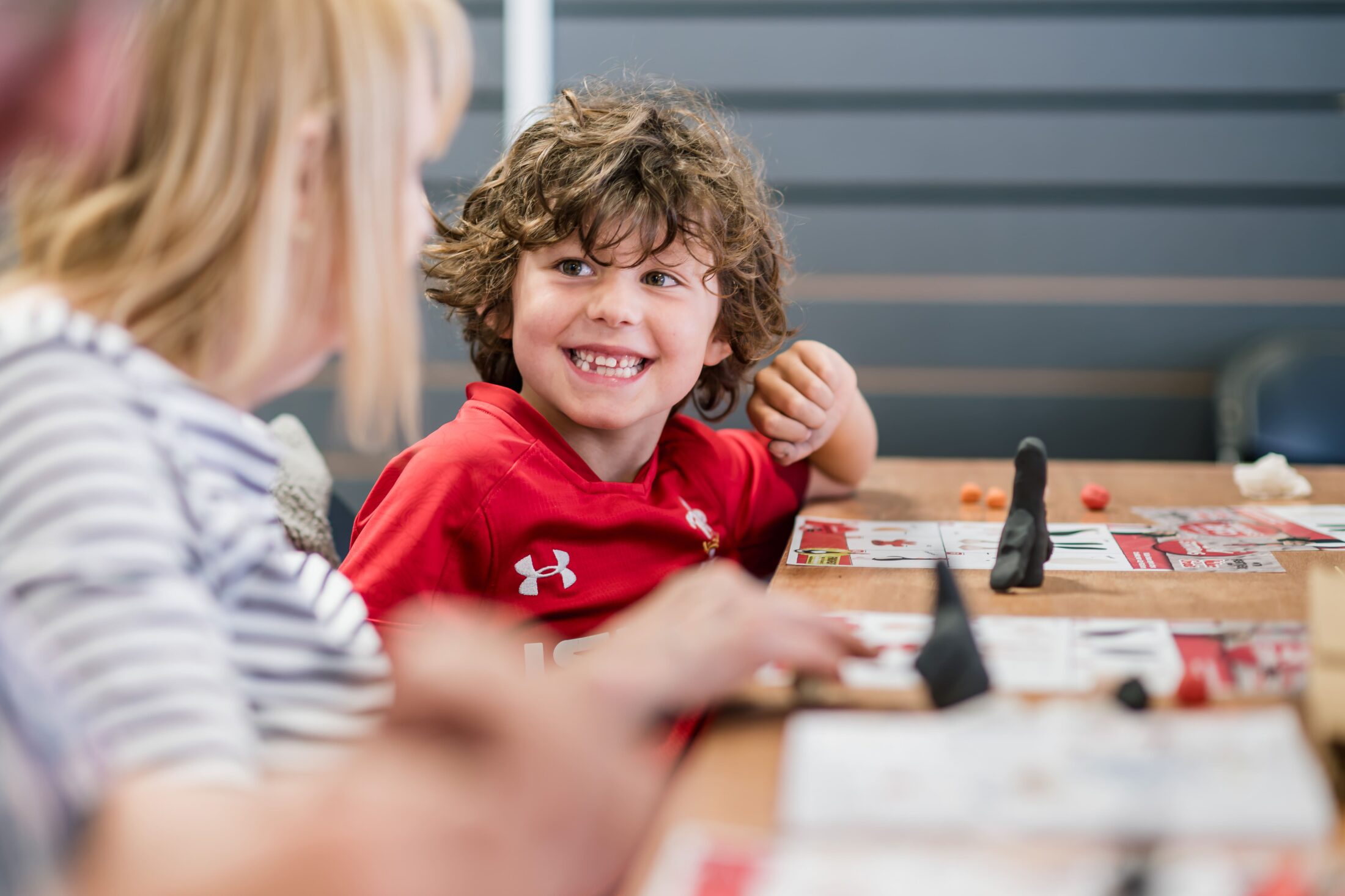 A young boy with curly hair smiles at an adult across a table while working on crafts. Art supplies and papers are spread out in front of them. The setting appears casual and friendly.
