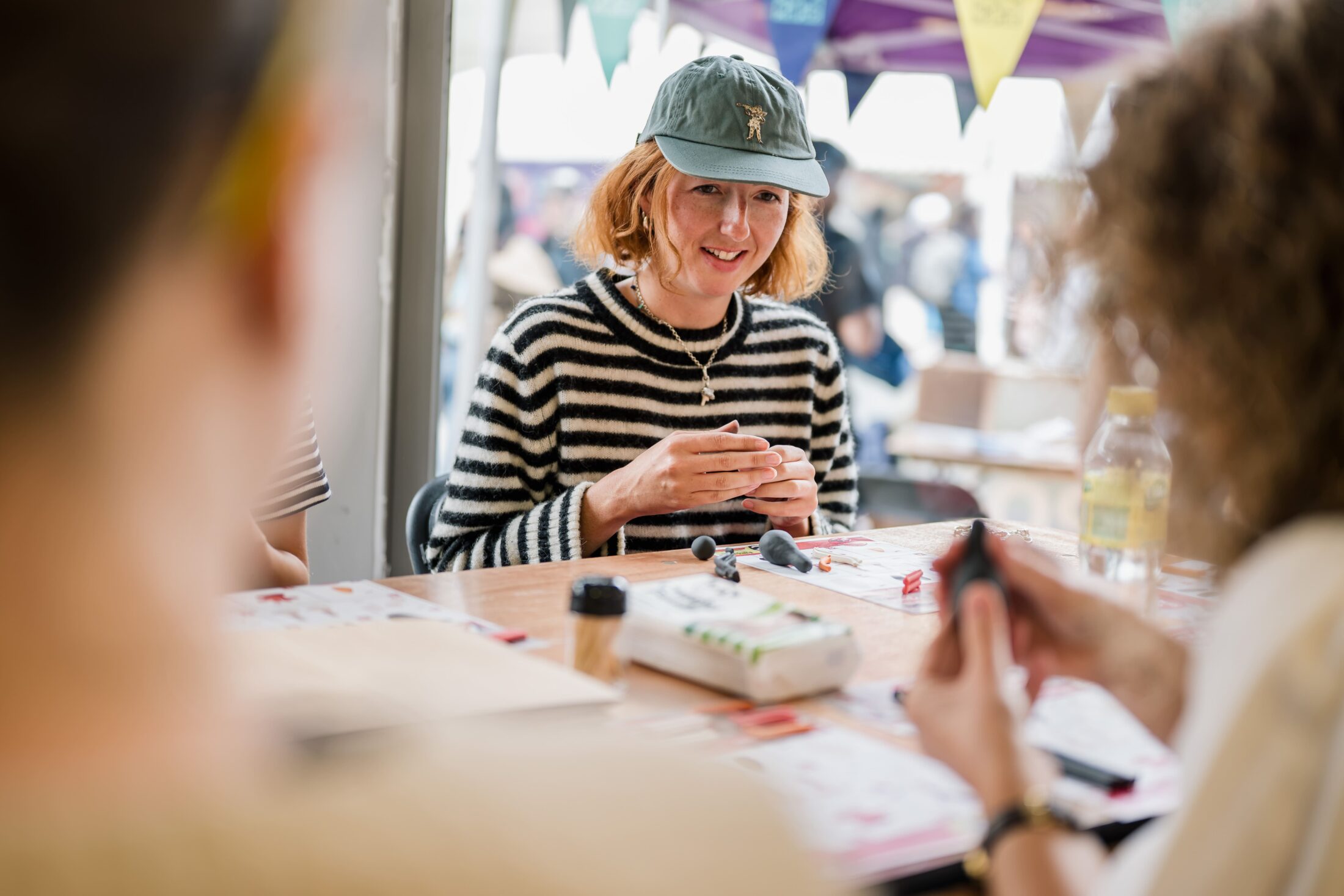 A person in a striped jumper and cap smiles whilst sitting at a table with art materials, participating in a creative activity with others at an outdoor event.