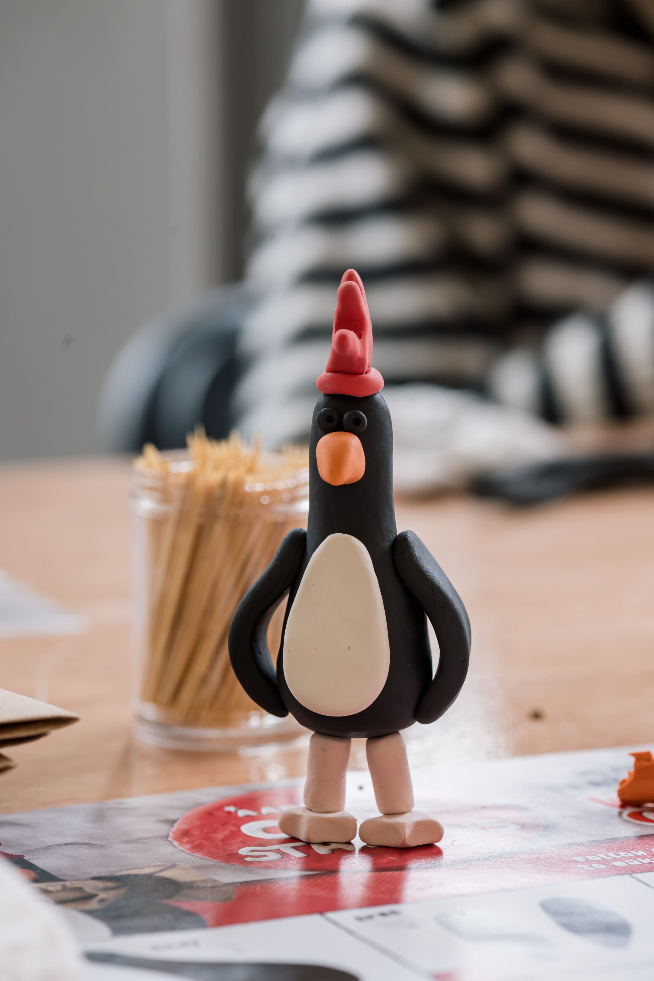 A clay penguin with a red hat stands on a table with magazines and cocktail sticks, while a person in a striped shirt is blurred in the background.