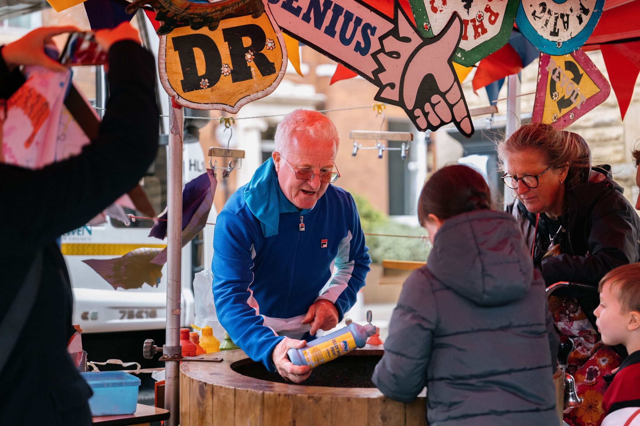 An older man in a blue jacket hands a bottle to a child at a colourful outdoor stall, while others watch. Bright banners and signs hang above, and a woman assists nearby.