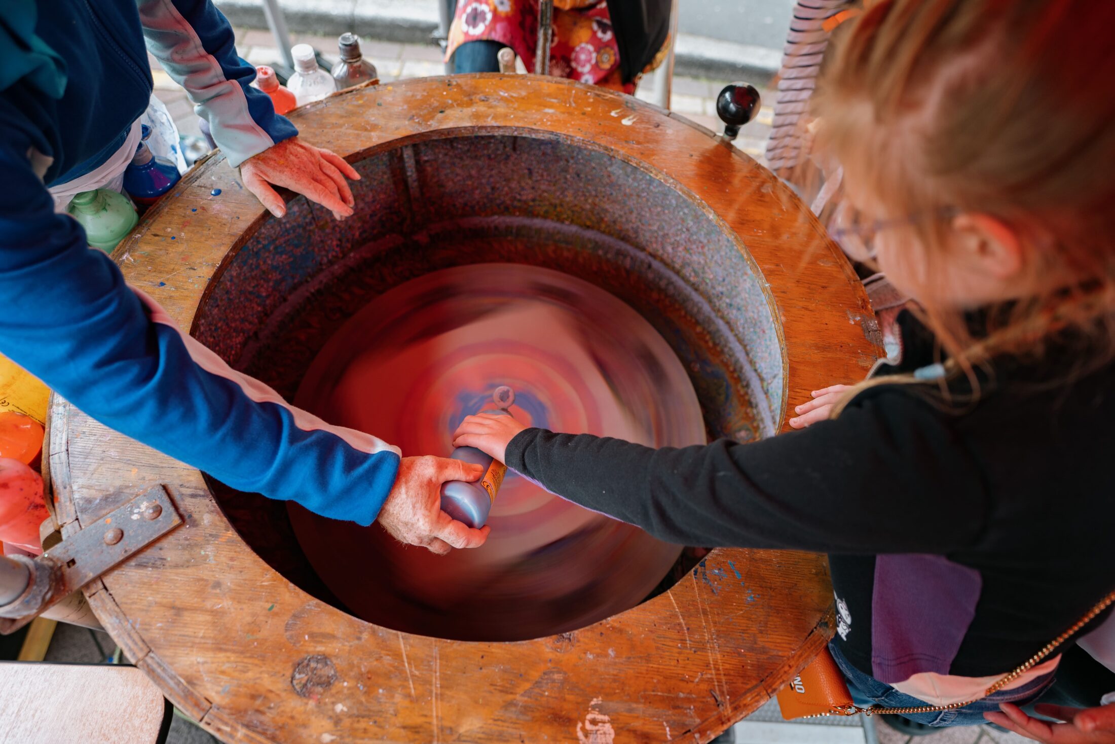 Two people swirl paint bottles over a spinning circular surface, creating colourful patterns in a large wooden bowl at an outdoor event.