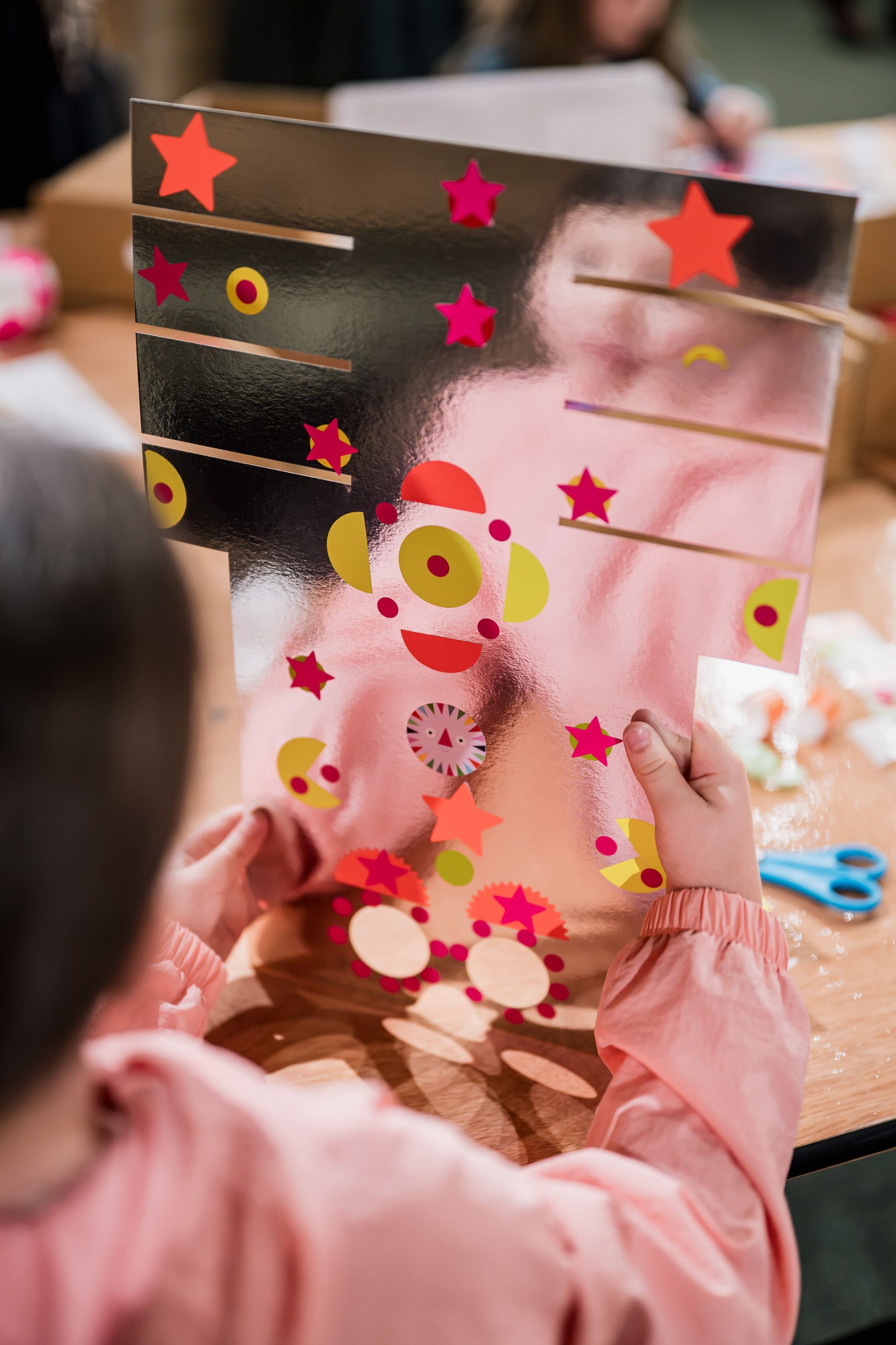 A child holds a shiny sheet decorated with colourful star and circle stickers at a table, with scissors and paper scraps nearby.