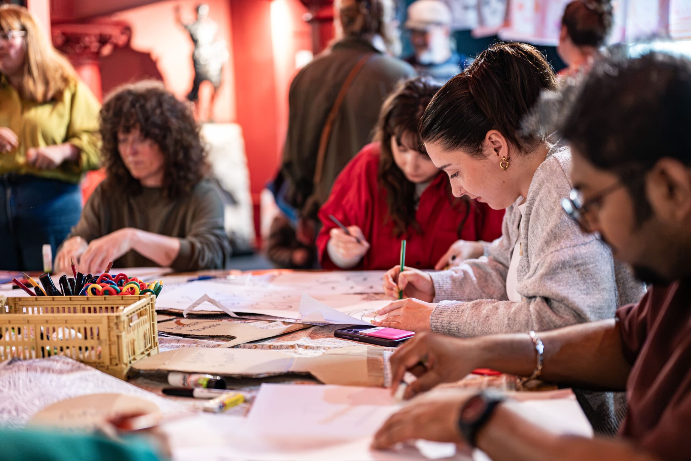 People sit around a large table, drawing and writing on paper with pens and markers. Art supplies are scattered on the table. The focus is on a woman in a grey jumper writing intently. The background is busy and colourful.