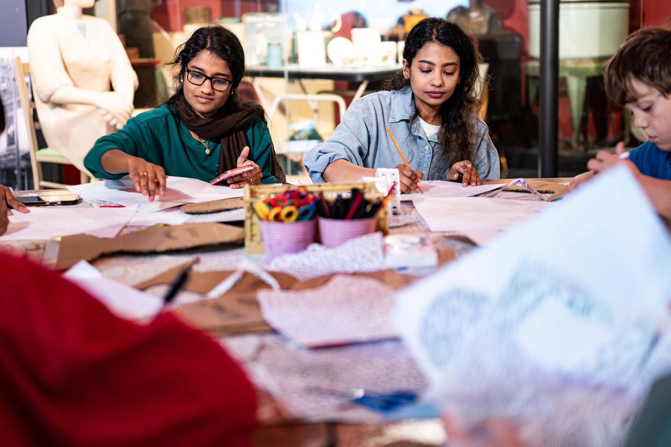 Two women sit at a table covered with art supplies and paper, focusing on drawing or crafting. Another person, partially visible, is also working at the table. The background shows shelves and mannequins.