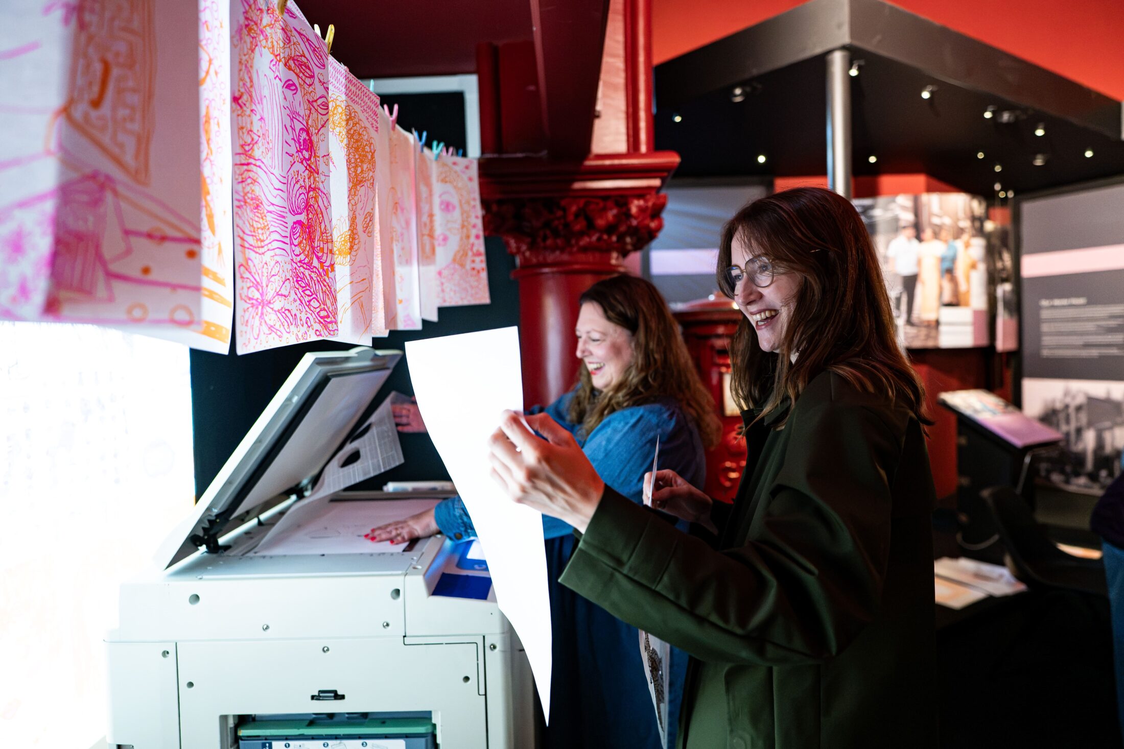Two women smile while using a photocopier in a colourful, modern exhibition space. Artworks hang above the copier under bright lights, and one woman holds a fresh printout.