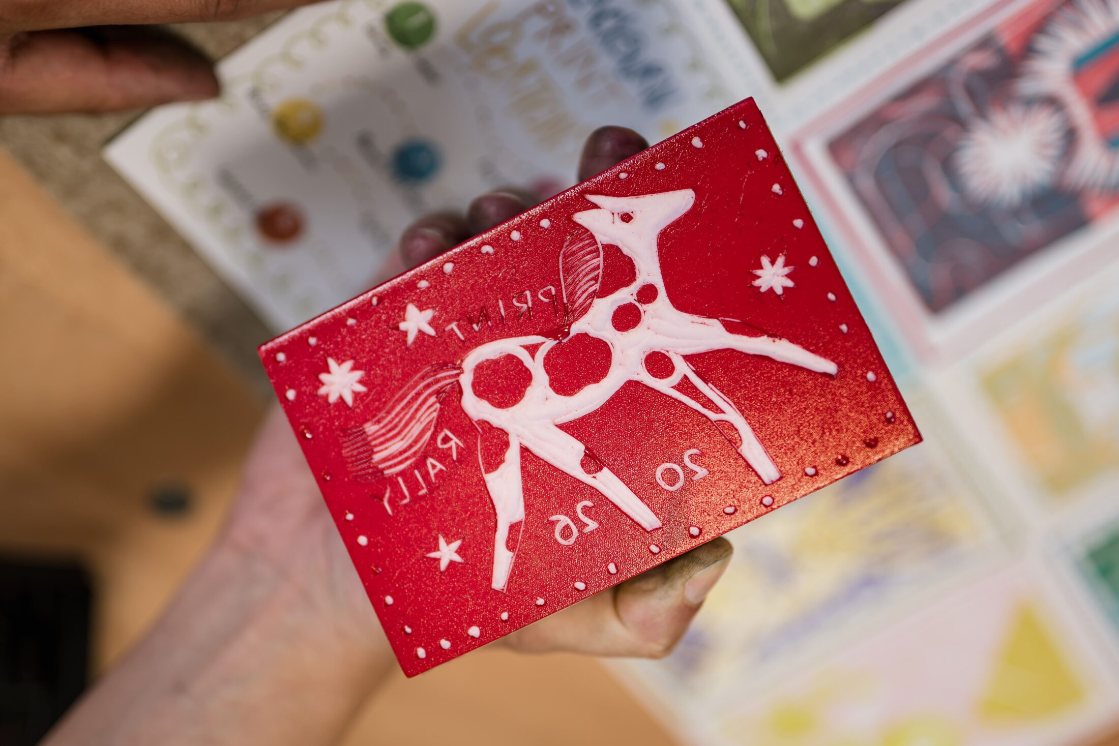 A hand holds a red card with a white stylised horse, stars, and the text Ralli 20 26. Colourful artwork or cards are blurred in the background.