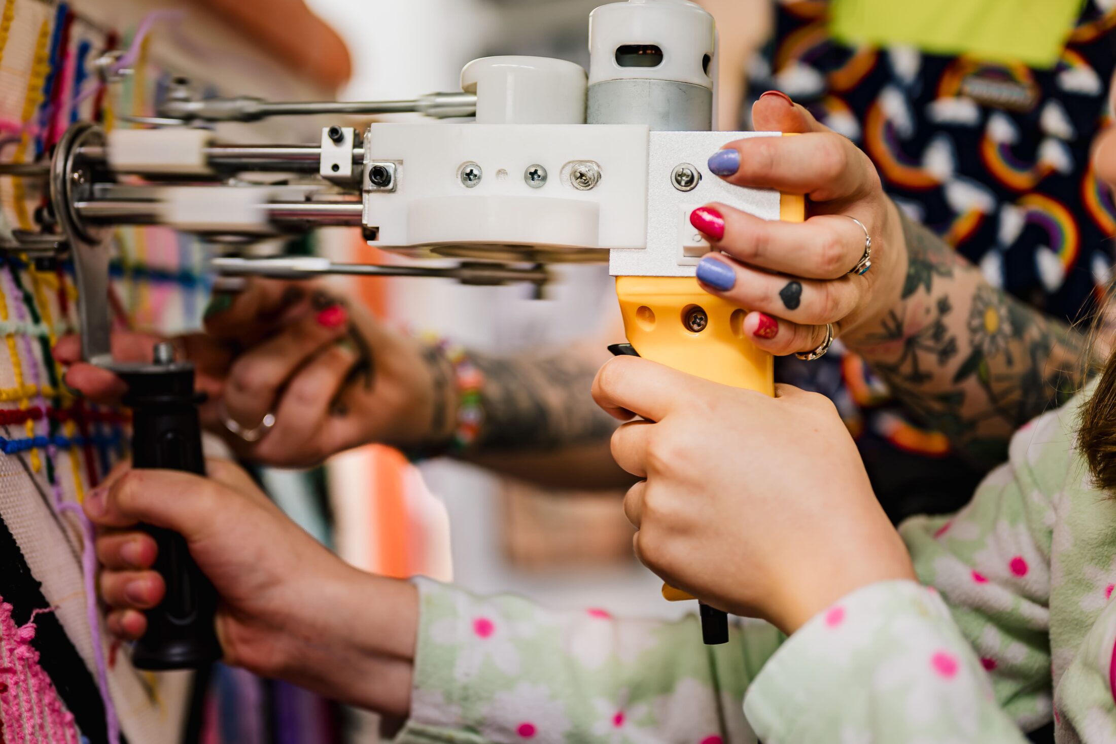 Two people use a yellow tufting gun on fabric; one guides the tool while another assists. Both have colourful manicures and tattoos. The background is blurred, showing patterned clothing and part of a textile artwork.