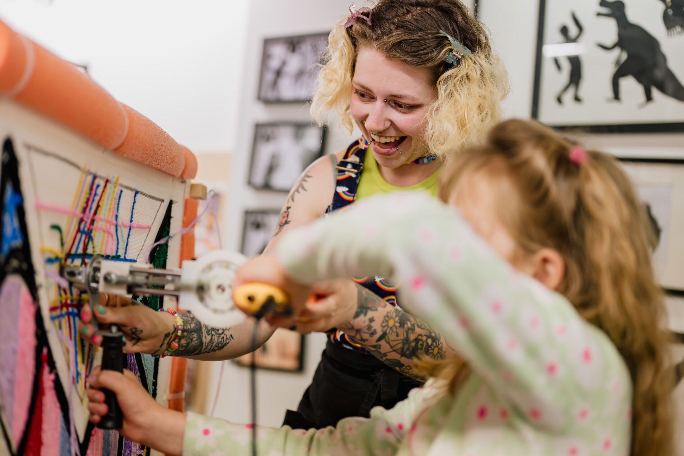 An adult and a child use a tufting gun to create textile art on a canvas frame, smiling and focusing on their work in a creative studio filled with framed artwork.
