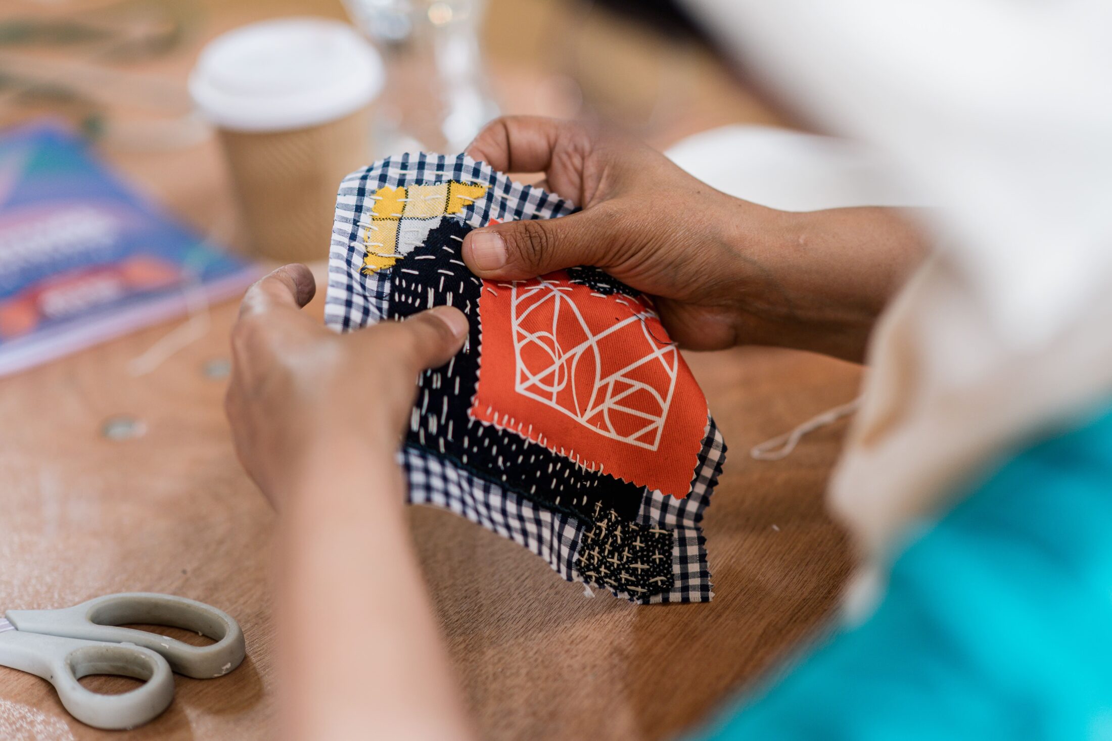 A person holds a piece of fabric with colourful patchwork and stitching. Scissors and a coffee cup are on the wooden table nearby.