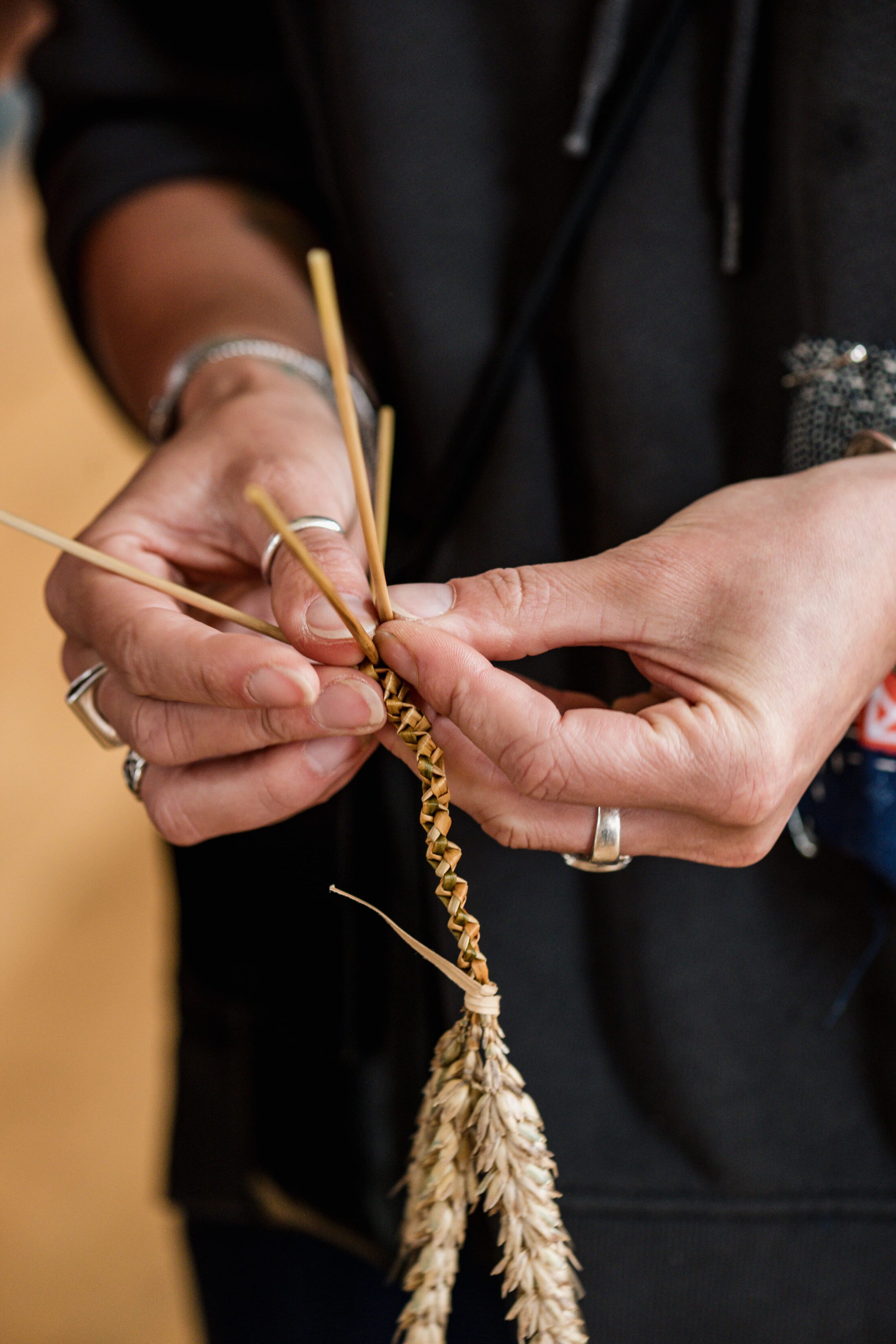 Close-up of hands plaiting dried wheat stalks together, creating a decorative braid. The person is wearing several silver rings and a dark long-sleeved shirt.