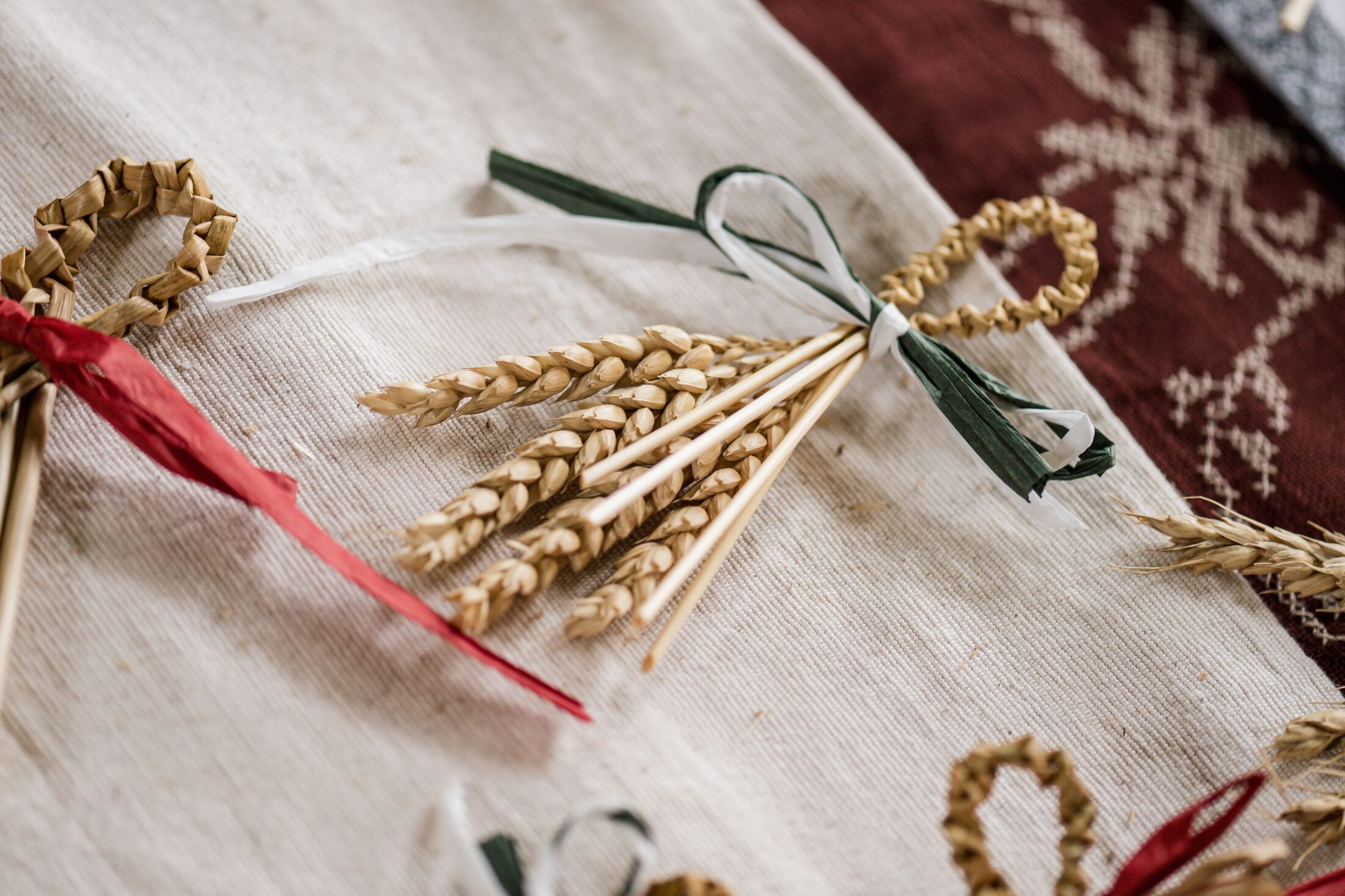 Close-up of handmade straw ornaments with wheat stalks tied together using coloured ribbons, arranged on a fabric surface with traditional patterns.
