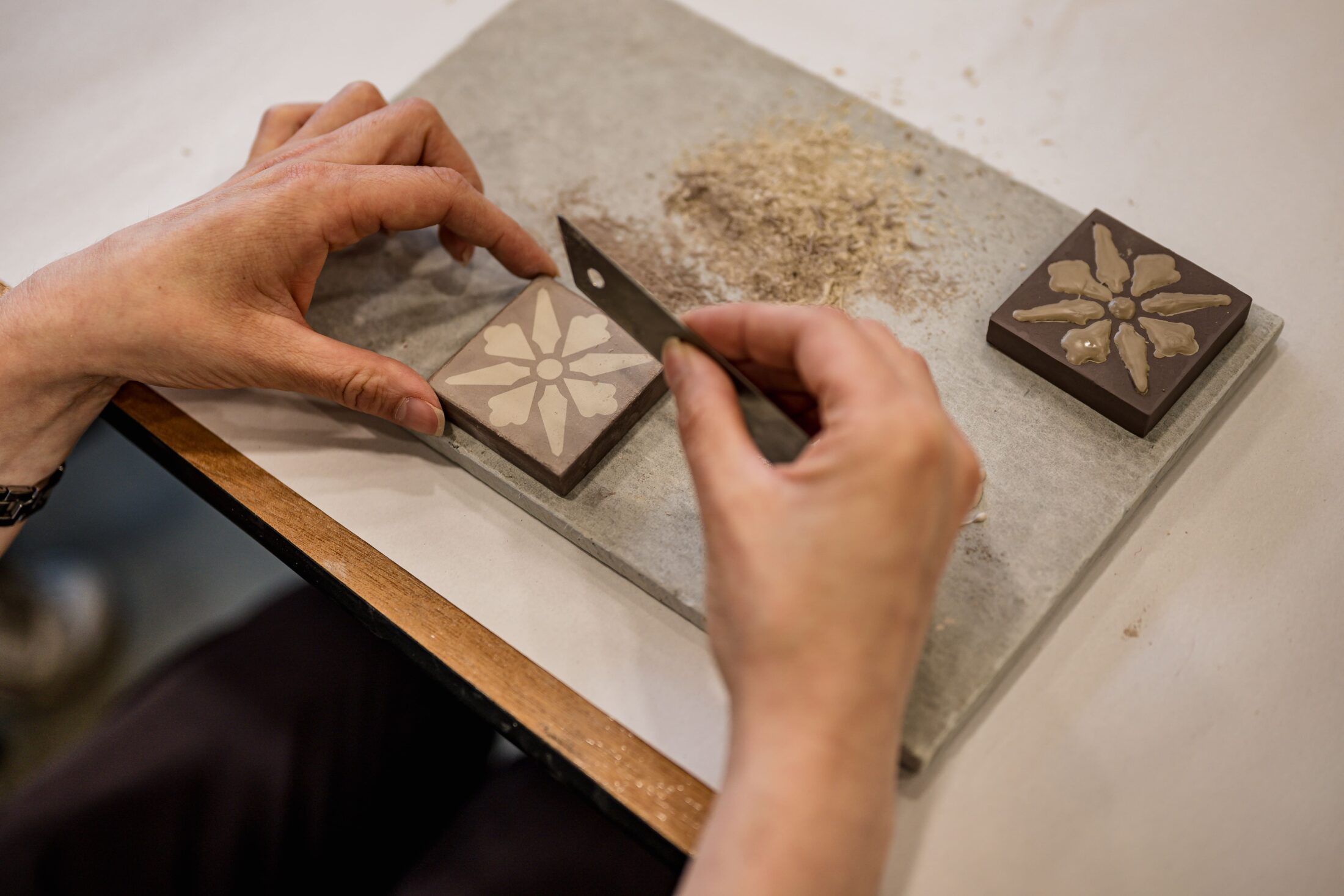A person uses a tool to carve a floral pattern into a square clay tile, with another finished tile and some dust on a white surface nearby.