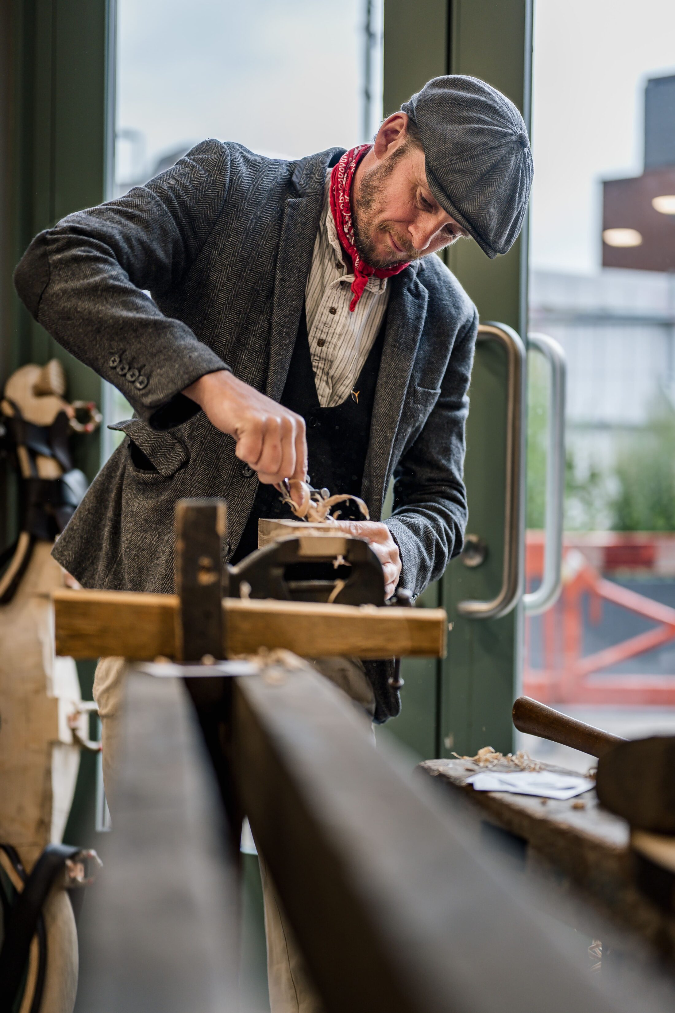 A man in vintage clothing and a flat cap uses a hand plane to shape wood indoors, standing by large windows with natural light streaming in.