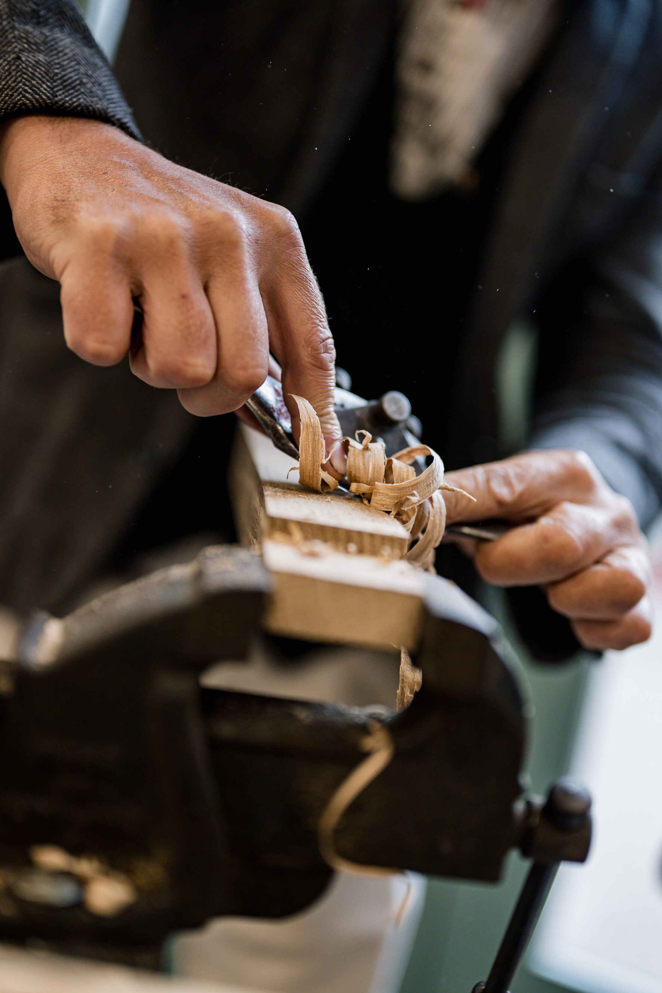 Close-up of hands carving wood with a chisel, creating curly wood shavings on a piece held in a vice.