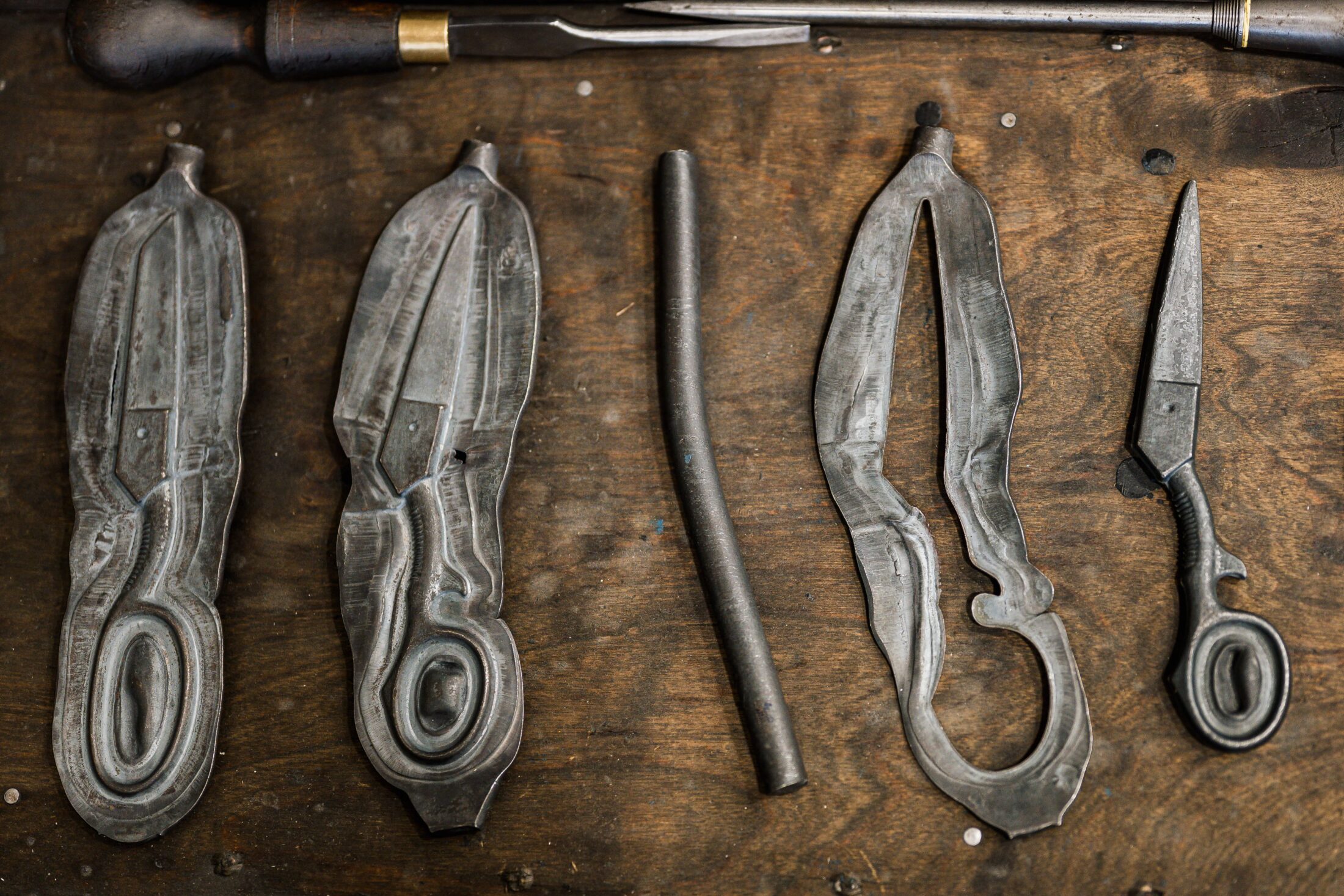 Four metal objects on a wooden surface: two mould halves shaped like scissors, a straight metal rod, an open mould, and a completed pair of metal scissors with a looped handle and pointed blade.