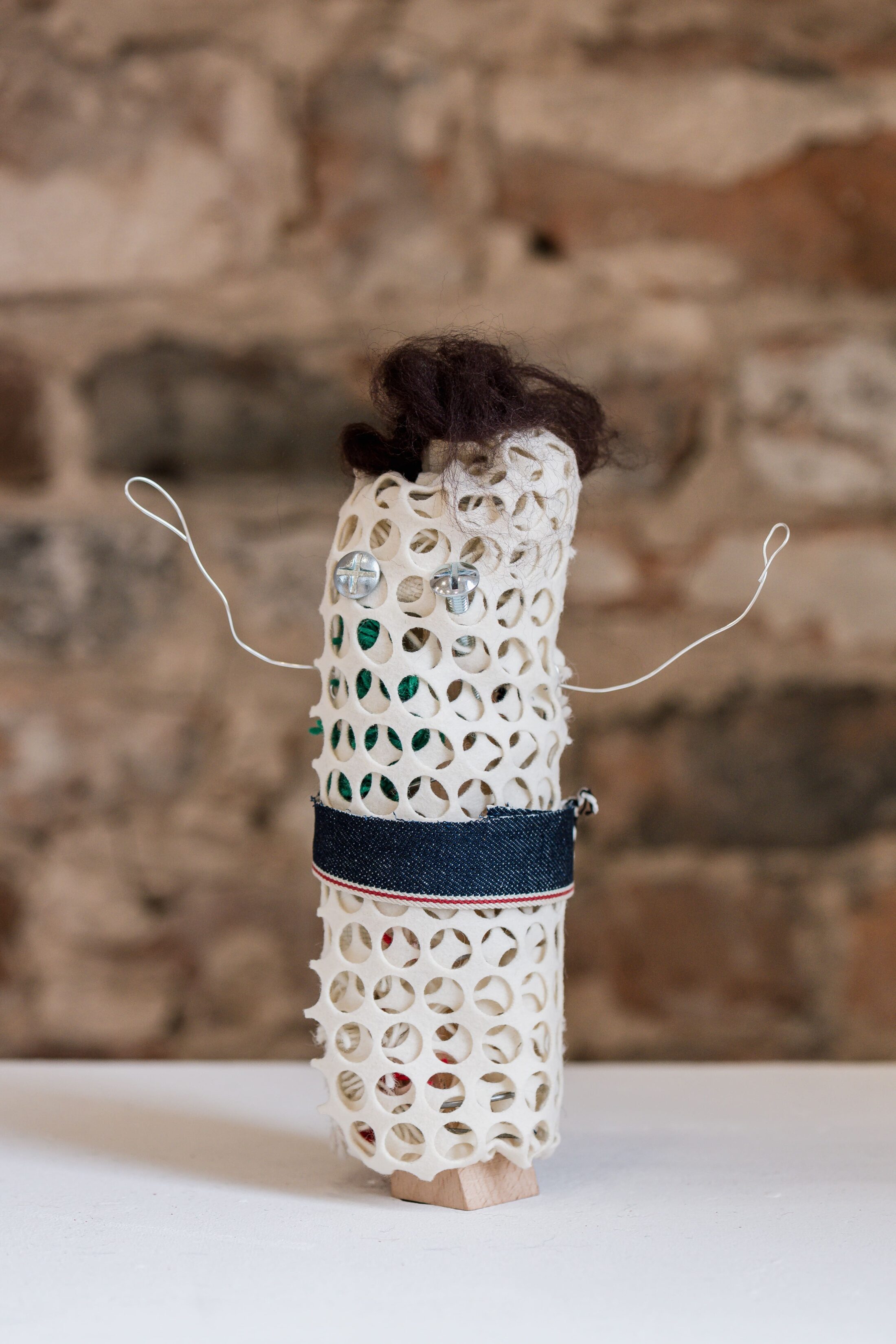A handmade figure with a mesh-like body, wire arms, a denim belt, small glass eyes, dark wool hair, and a stone wall background.