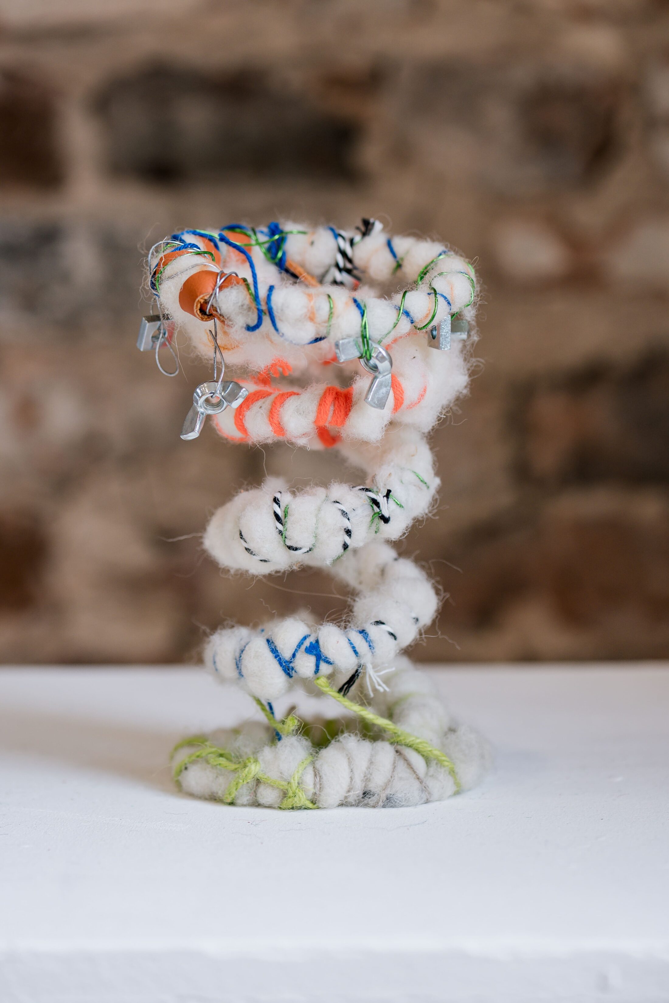 A spiral sculpture made of fluffy, white material with coloured threads and small metal clips, displayed on a white surface with a blurred stone wall background.