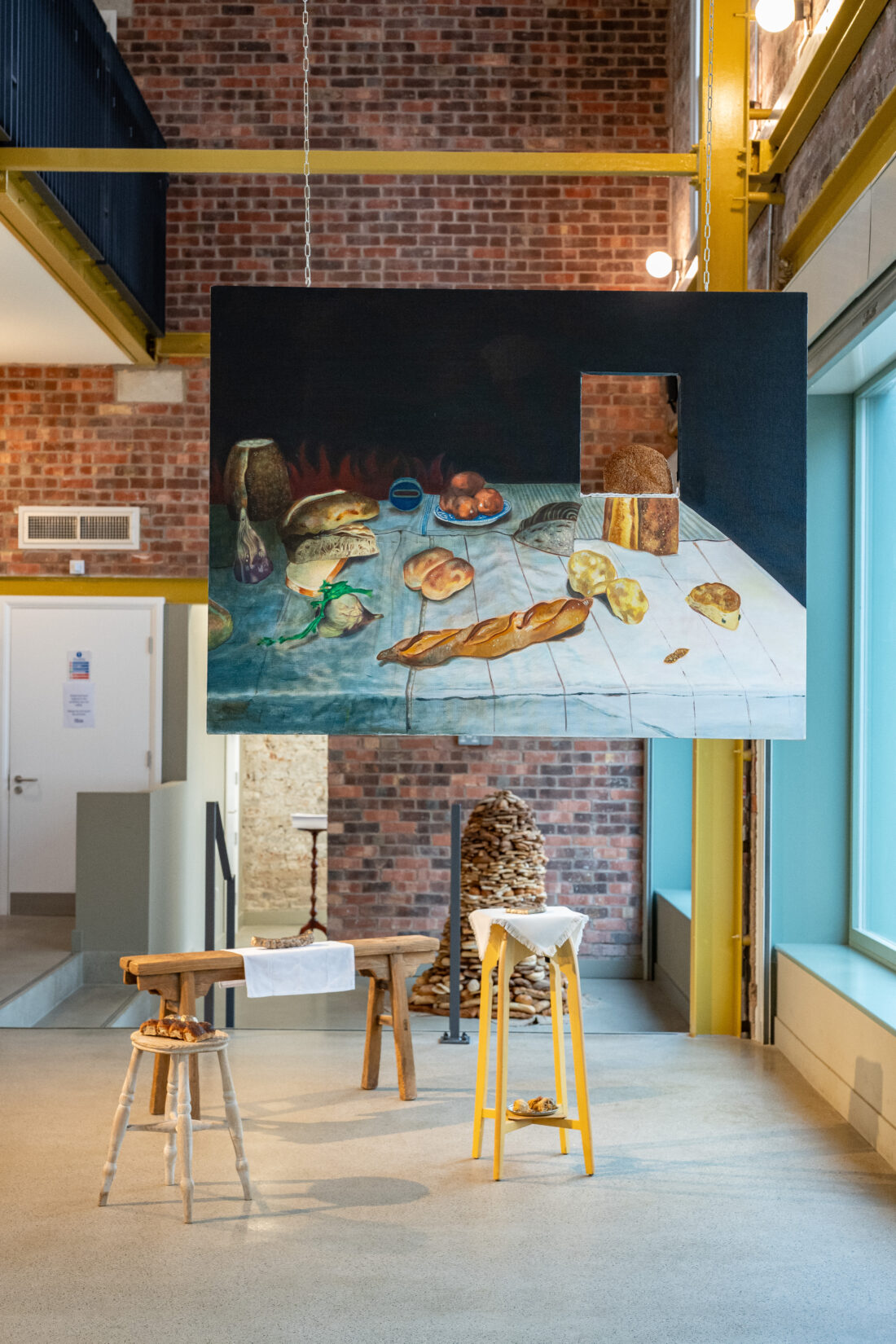 A painting of bread and produce hangs in a modern gallery. Below, tables and stools display real bread loaves, echoing elements from the artwork. Brick walls and large windows frame the bright, contemporary space.