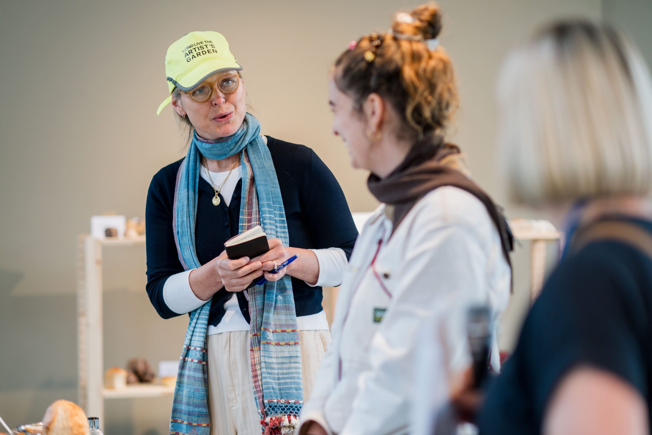 A woman in a yellow hat and blue scarf takes notes whilst talking to two other women, one in a white coat, inside a brightly lit room with shelves in the background.