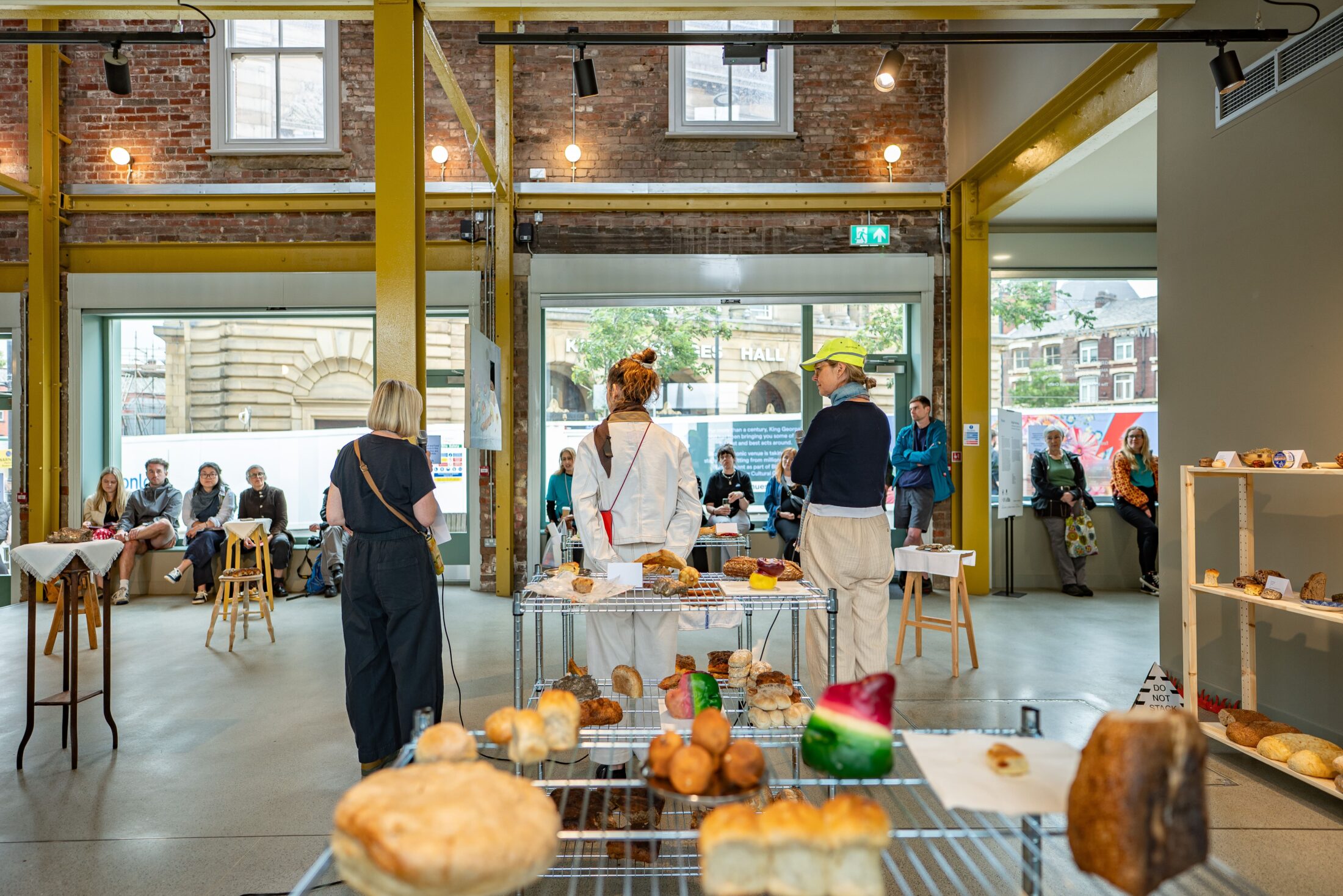 Several people stand around tables with bread and pastries in a bright, industrial-style room, whilst others sit and watch near large windows. Shelves with more baked goods are on the right.