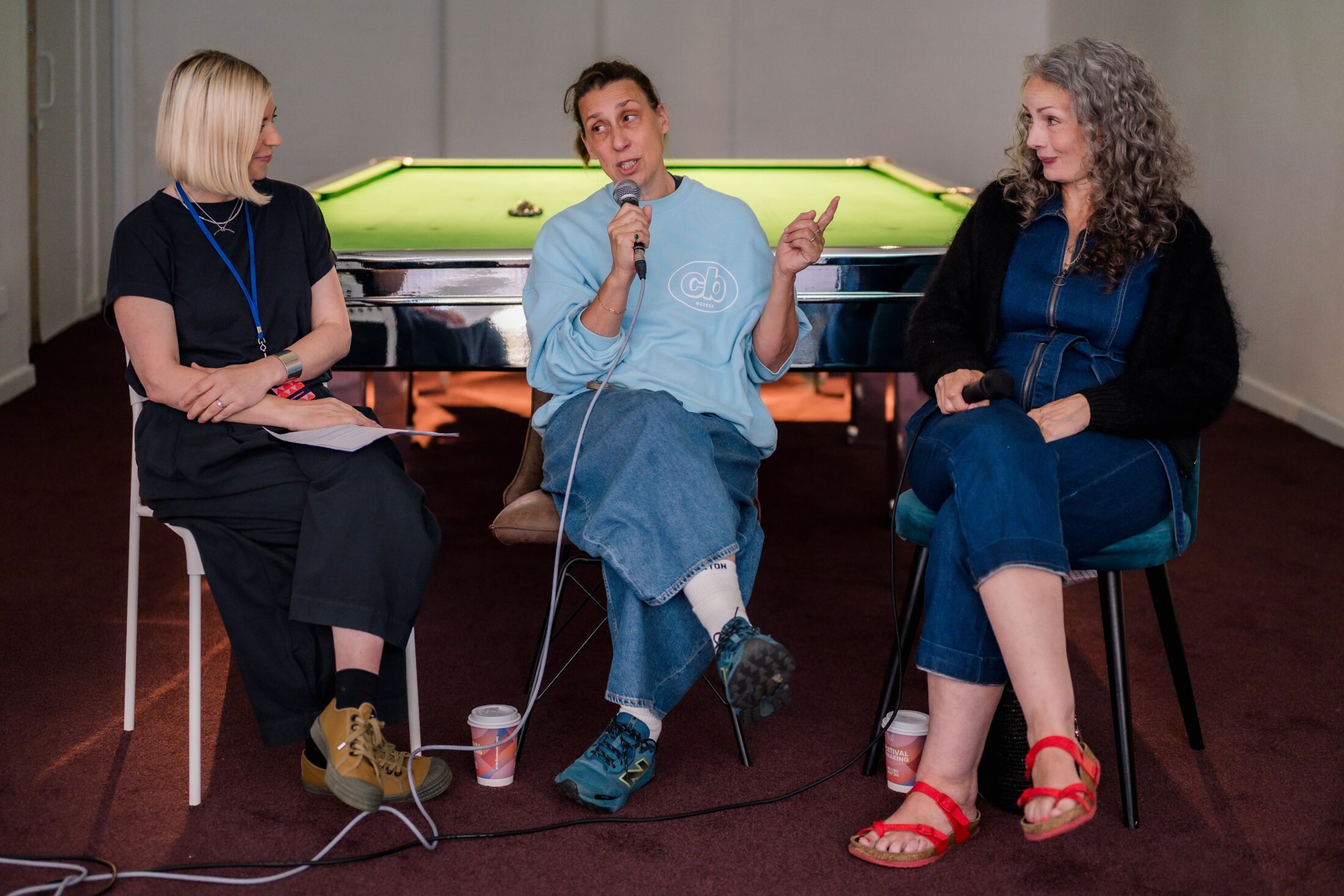 Three women sit in front of a pool table, talking. The woman in the middle speaks into a microphone whilst gesturing. The women on either side listen attentively. Disposable cups rest on the floor.