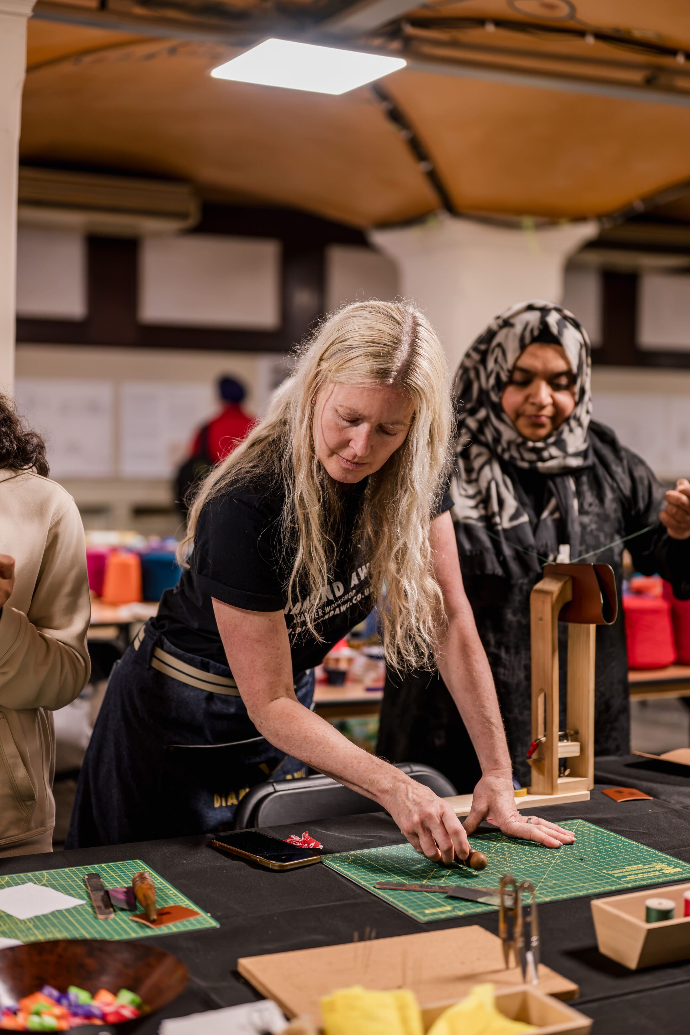 Two women work together at a crafting table, handling materials and tools. One woman, with long blonde hair, leans over the table, whilst the other, in a patterned headscarf, observes closely.