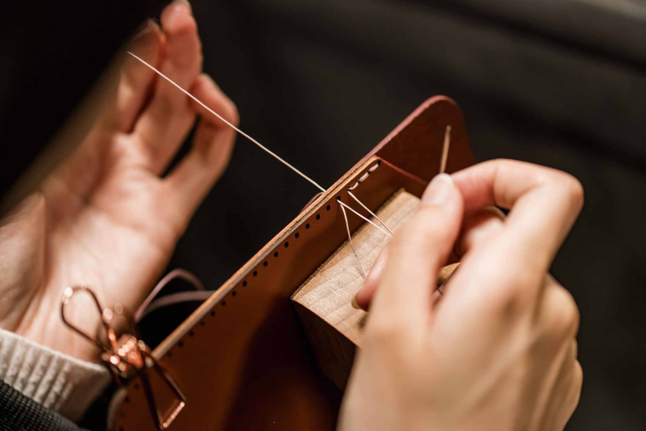 Close-up of hands stitching leather with a needle and thread, using a wooden stitching clamp to hold the material in place.