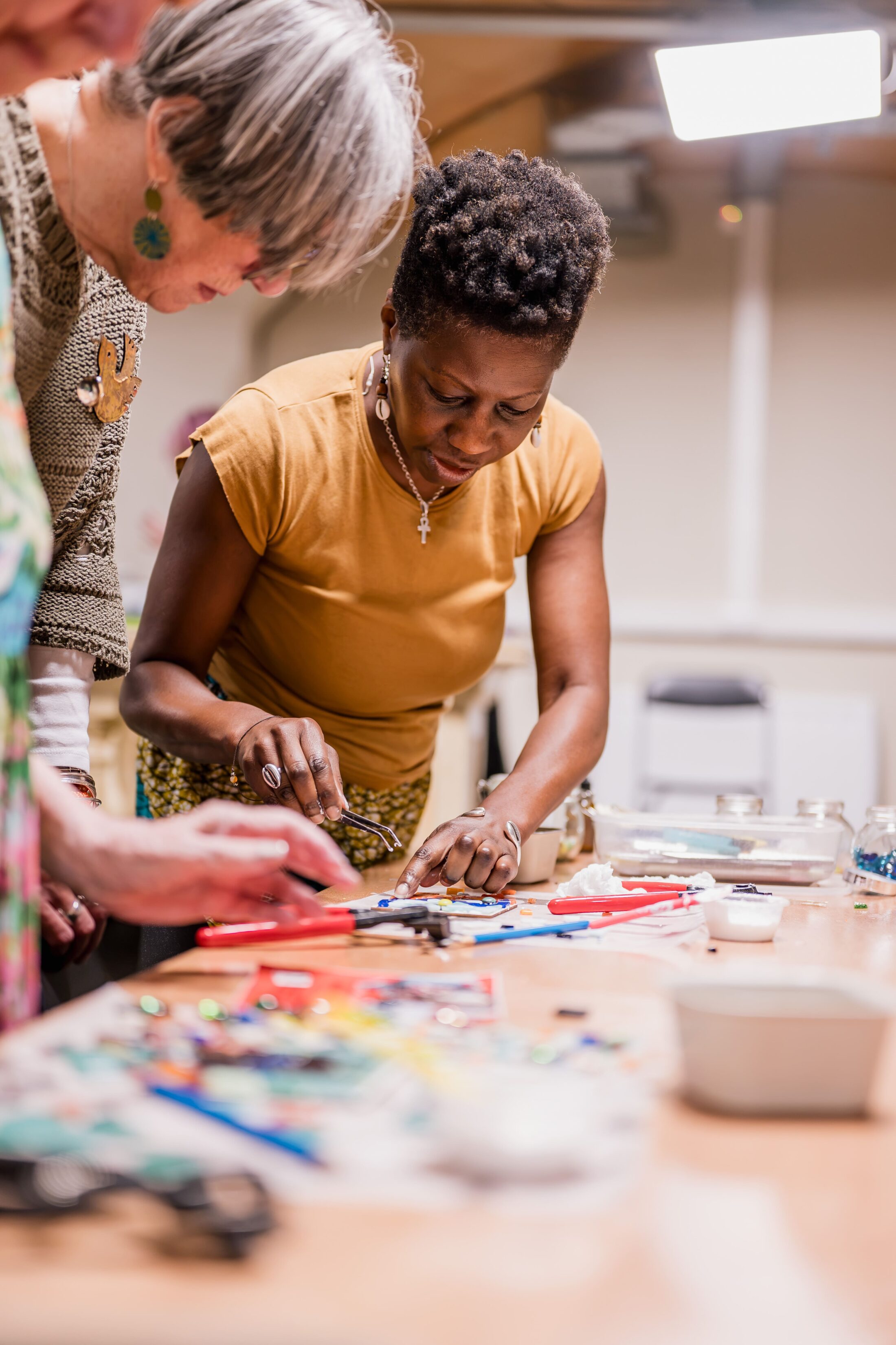 Three women lean over a table covered with colourful art supplies, focusing on a hands-on creative project together in a well-lit room.