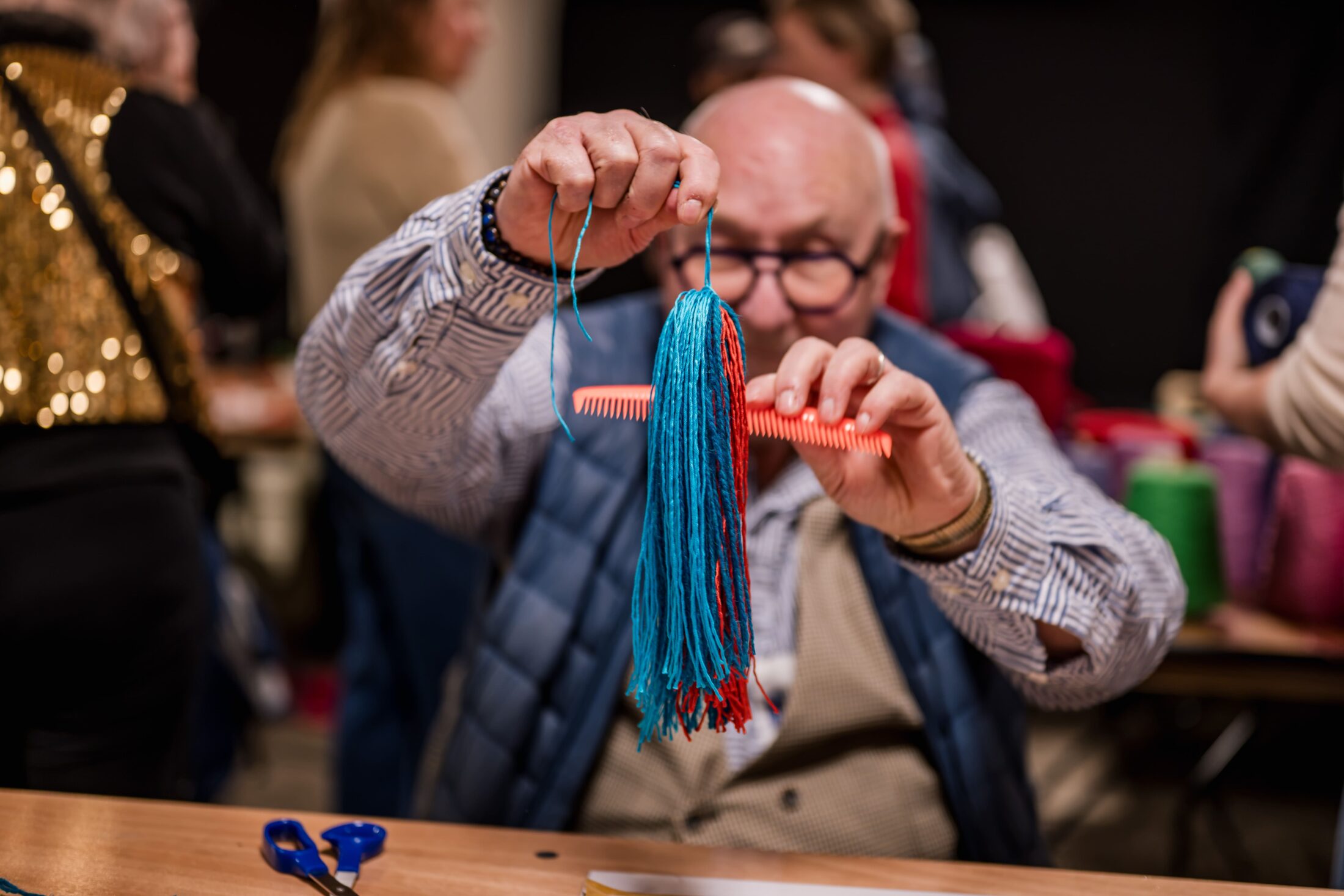 An older person wearing glasses holds up blue wool and a red comb, working on a craft project at a table. Other people are visible blurred in the background.