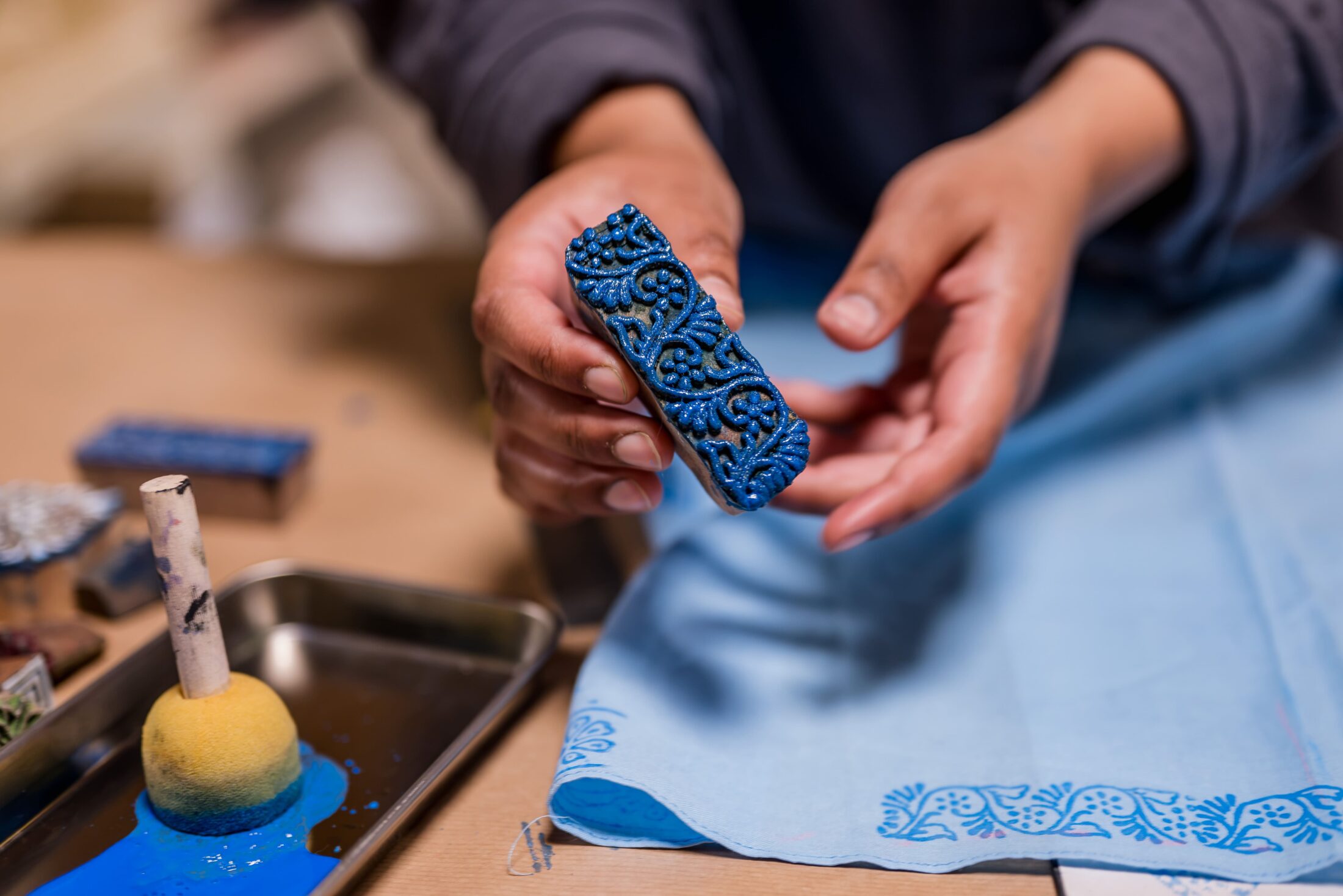 A person holds a small, blue, intricately carved wooden stamp above a piece of light blue fabric, with stamping tools and blue paint visible nearby.